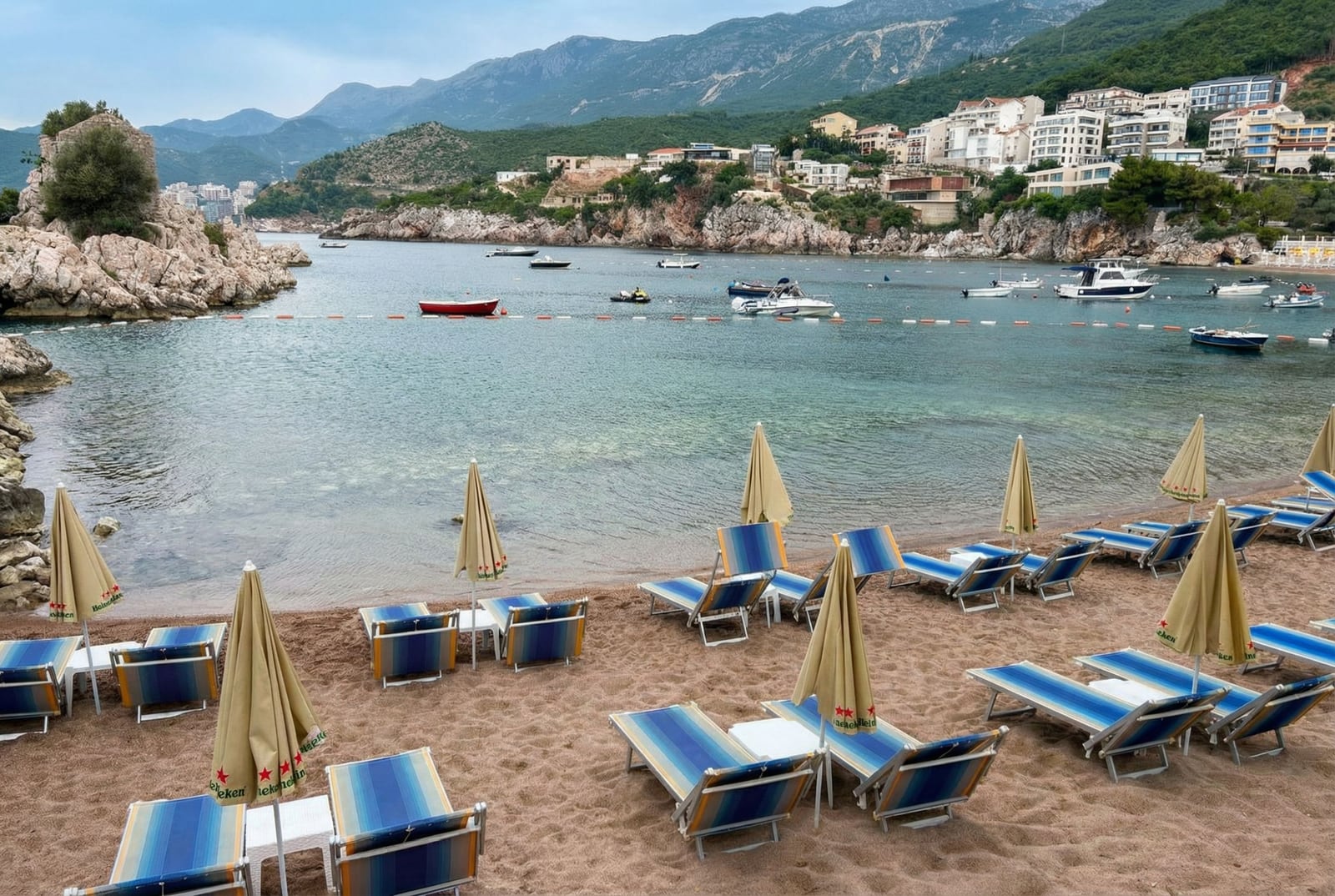 Sandy beach with sun loungers and umbrellas in Pržno, Montenegro, overlooking a picturesque bay, cliffs, and moored boats.