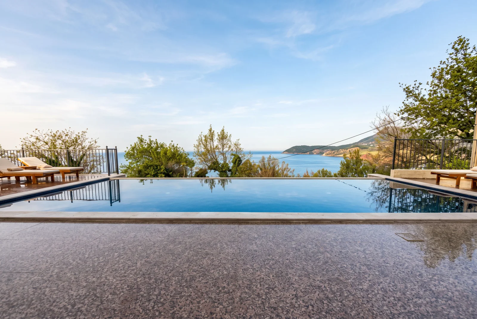 Infinity pool overlooking the sea and hills, wooden deck with sun loungers and metal railing under blue sky.