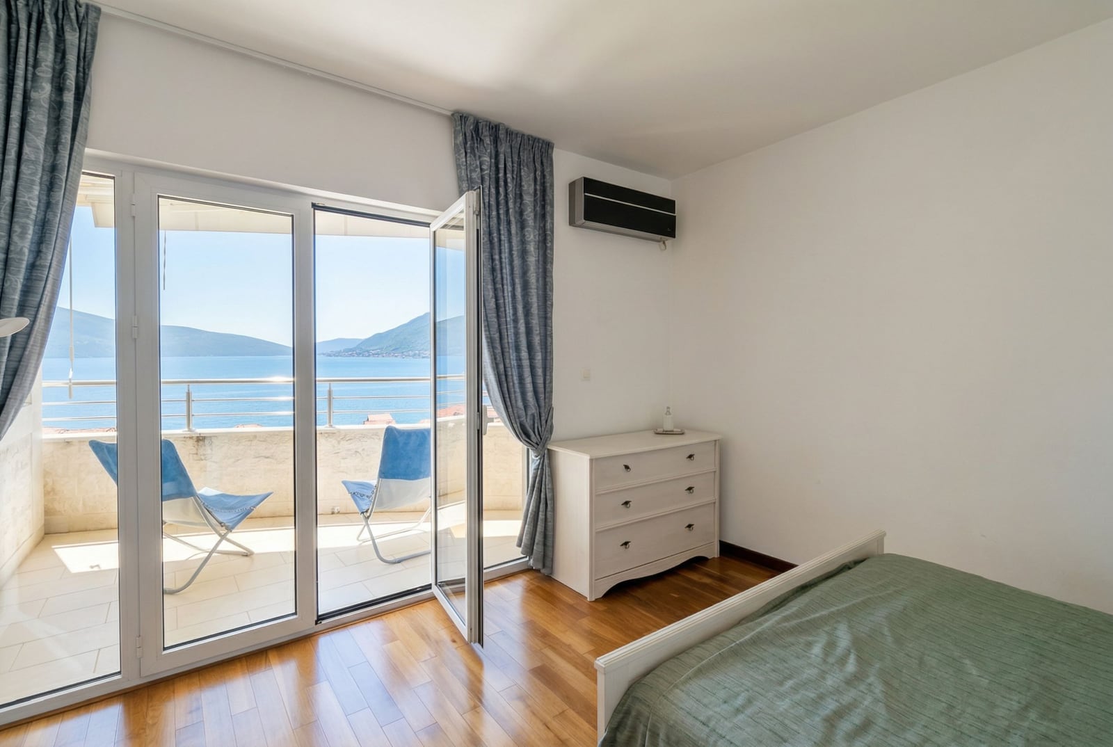 Bedroom in an apartment in Tivat, Montenegro: sea view from the balcony, light-colored furniture, parquet flooring, and panoramic windows.