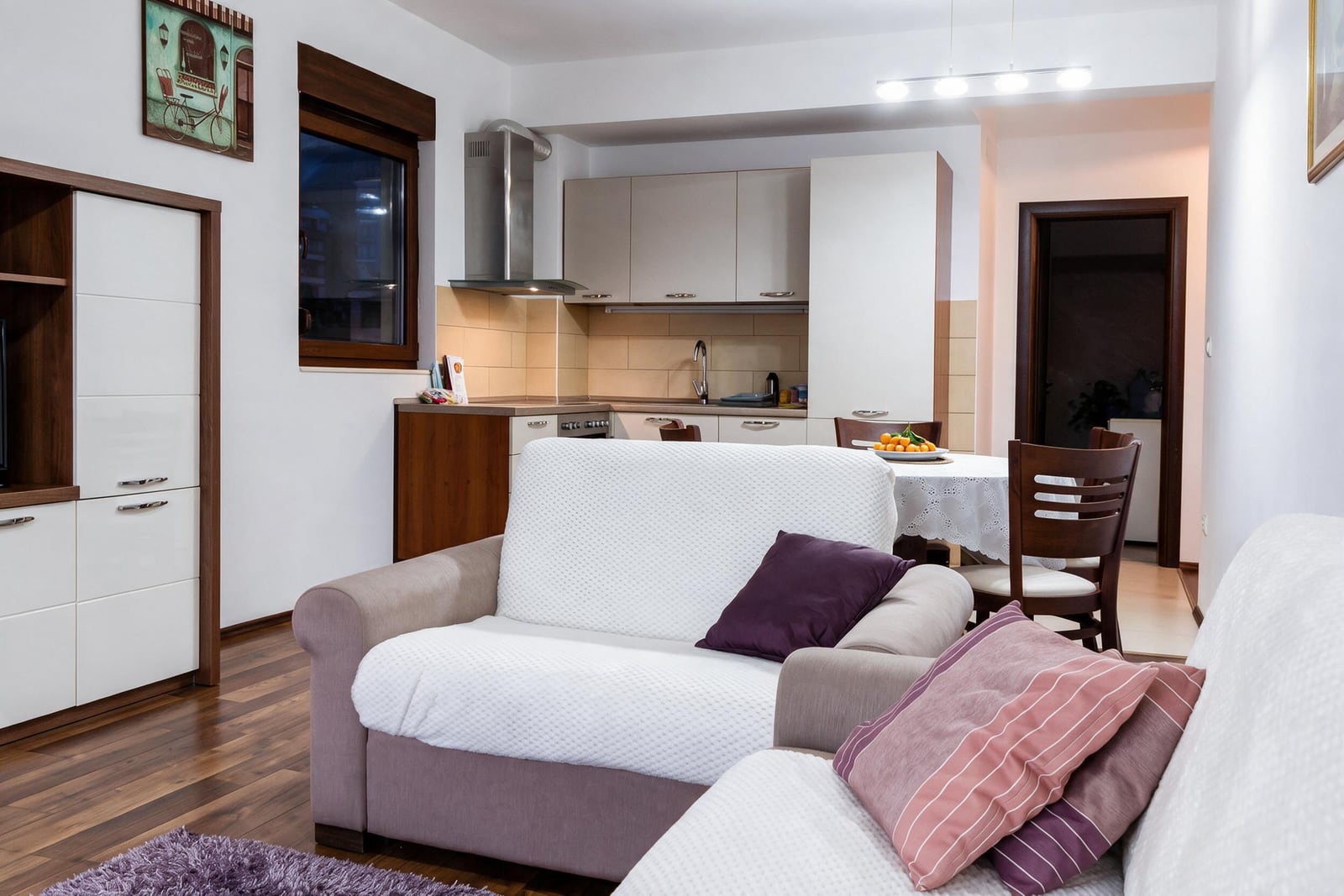Living room with sofa and open kitchen in background, wooden floor and storage unit.