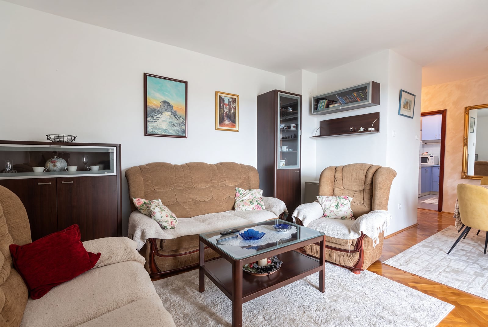 Living room featuring beige sofas, a glass coffee table, display cabinet, and framed paintings on white walls.