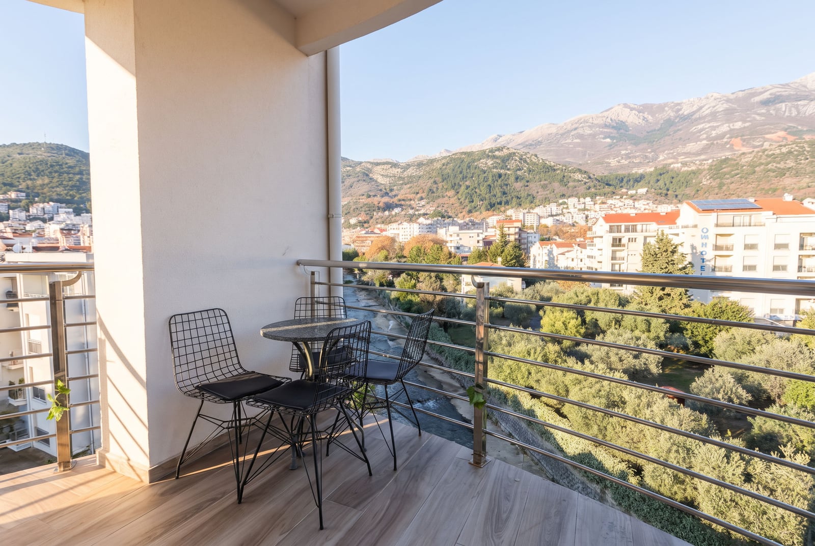 Balcony with black wire chairs and table overlooking a river, city, and mountains.