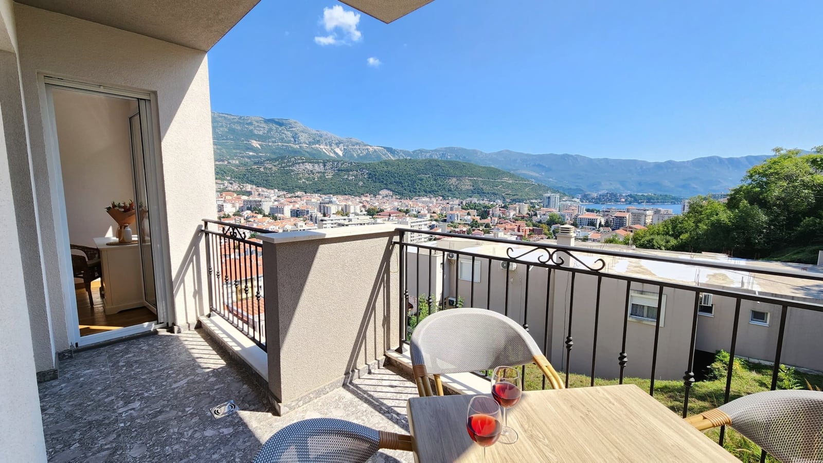 Balcony with a view of the city and mountains in a Budva apartment, Montenegro: a small table with wine glasses and wicker furniture.