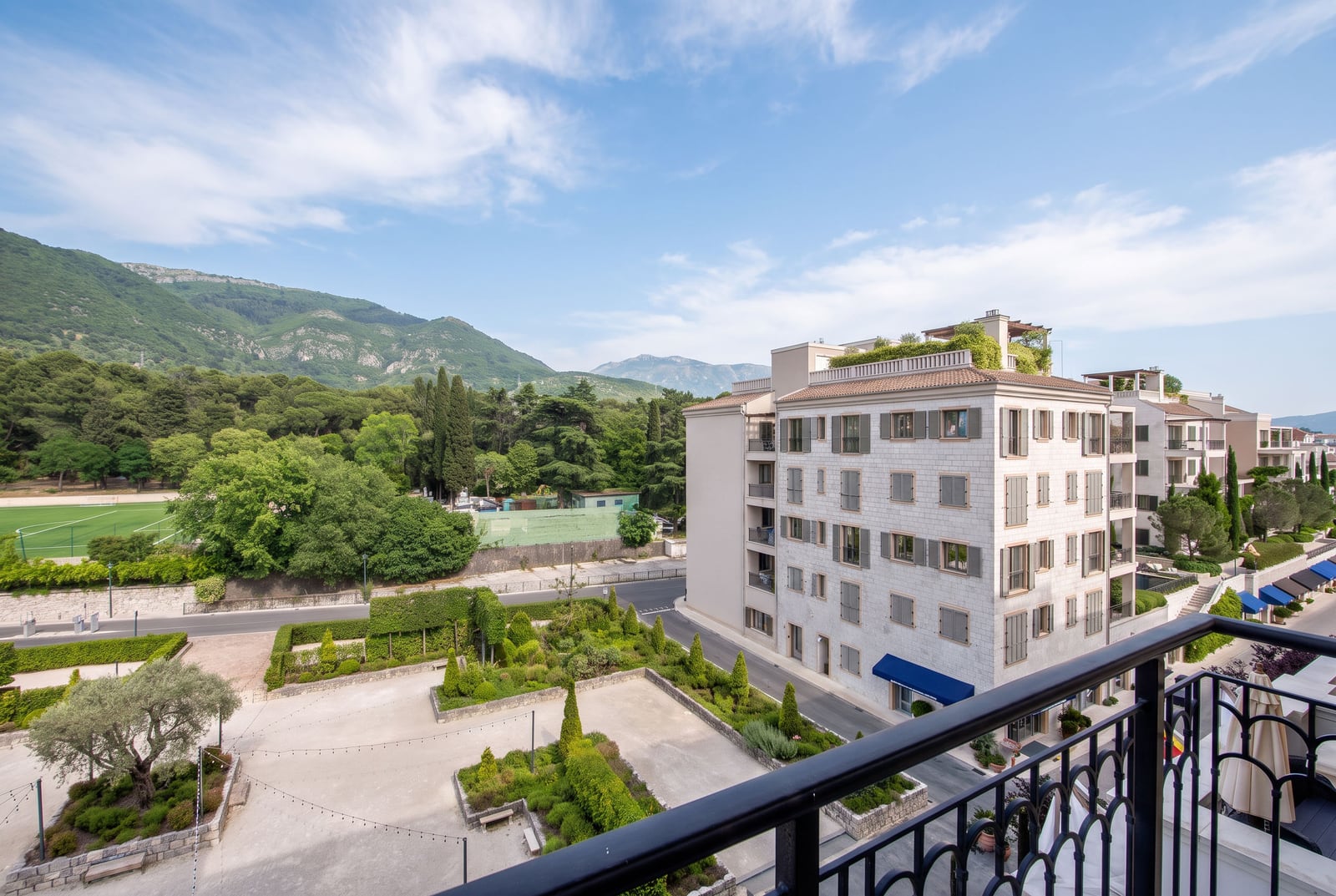 View from a balcony overlooking a garden, building, and mountains. Black iron railing is in the foreground.