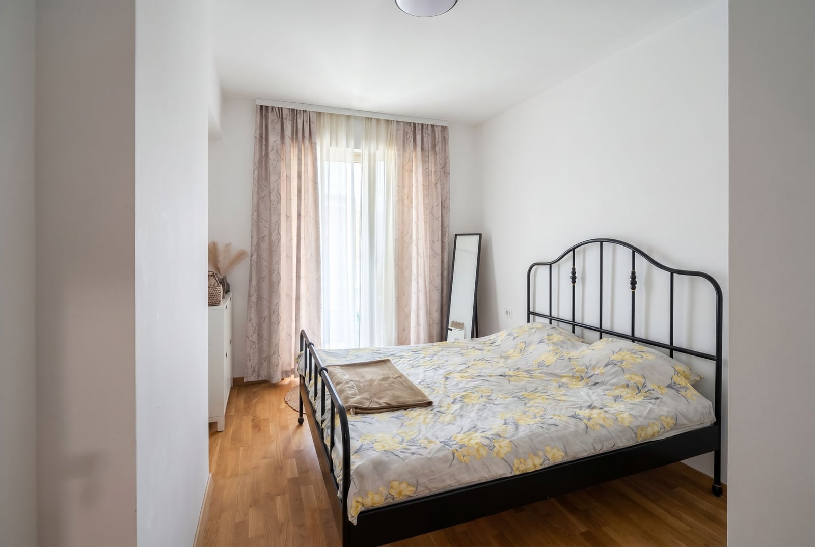 Bright bedroom with a black wrought-iron bed in an apartment in Becici, Montenegro: parquet flooring, panoramic window with curtains.