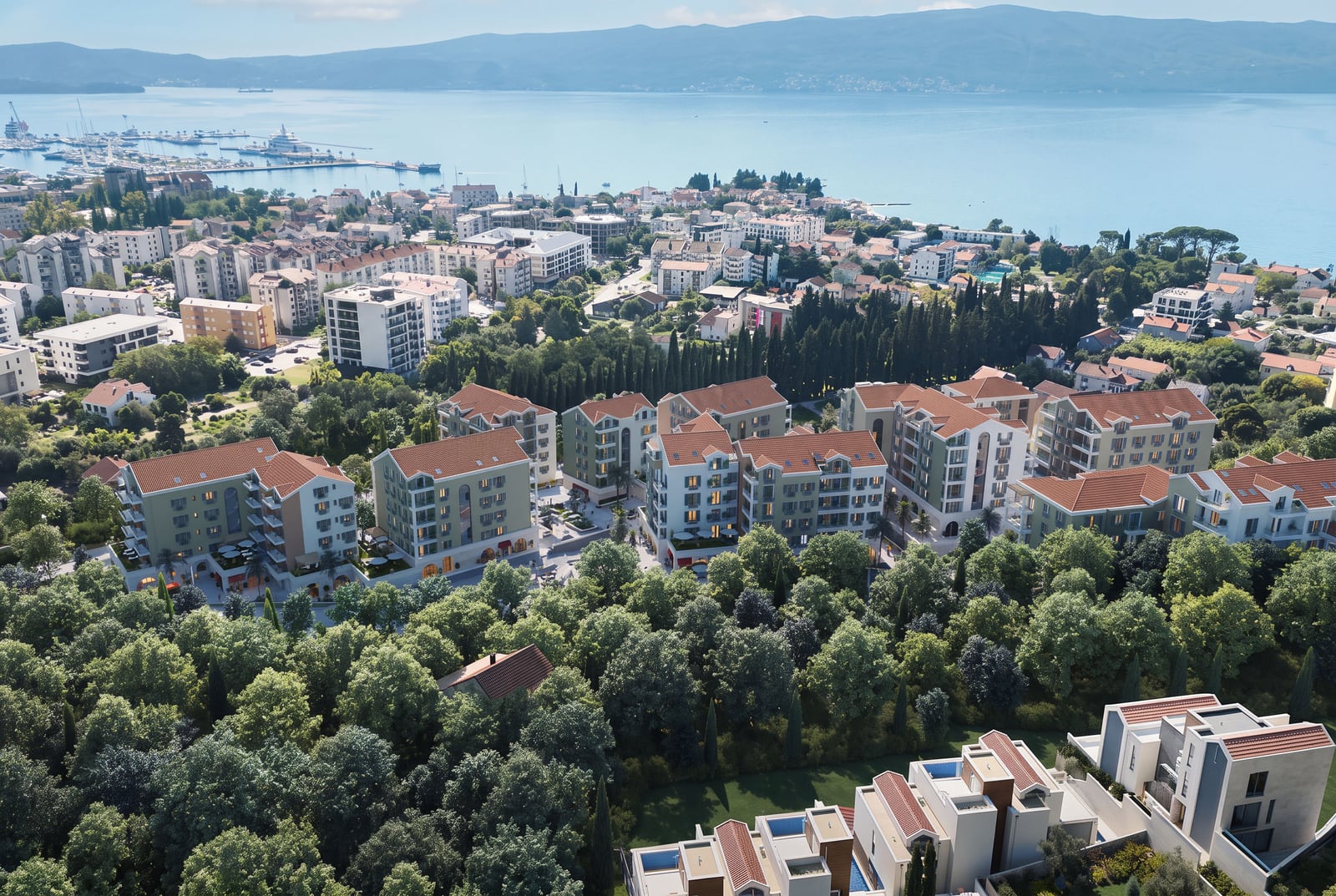 Aerial view of residential buildings with red roofs surrounded by trees, overlooking the sea and city.