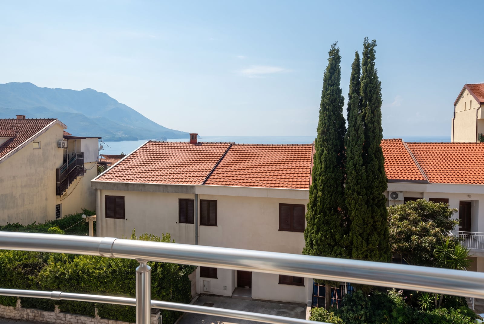 View from the balcony of an apartment in Bečići, Montenegro: tiled rooftops, green cypresses, mountains, and the sea.