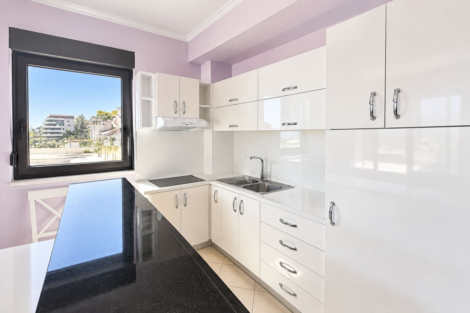 Modern kitchen with glossy white cabinets, black countertop, sink, cooktop, and a window overlooking buildings.