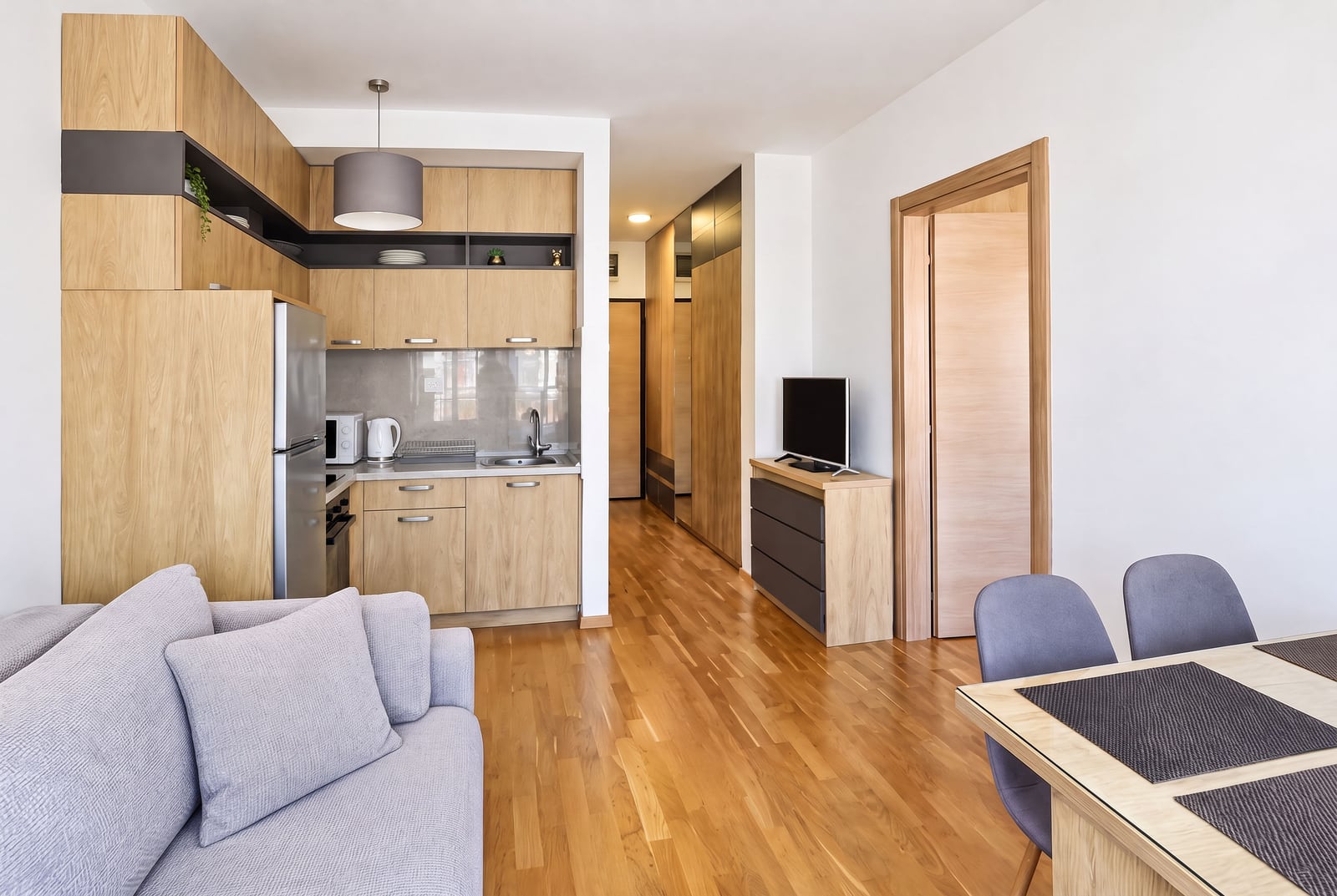 Living room with kitchen area, grey sofa, dining table, and wooden flooring.