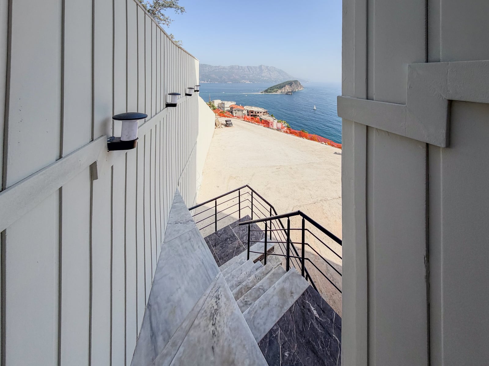 Marble staircase with black railing leads to an open terrace overlooking the sea and mountains.