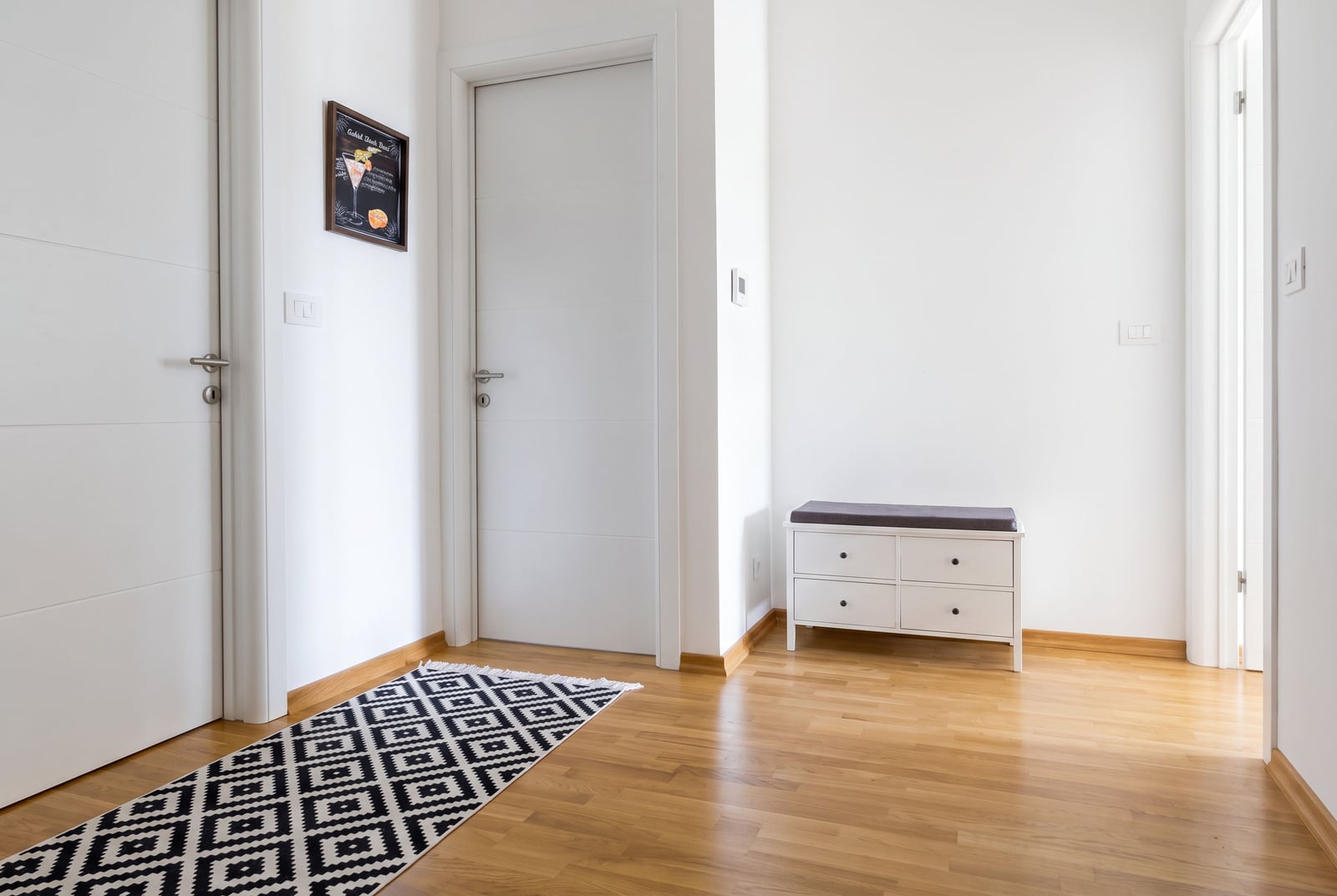 White hallway with wooden floor, patterned runner rug, storage bench, and framed picture on the wall.