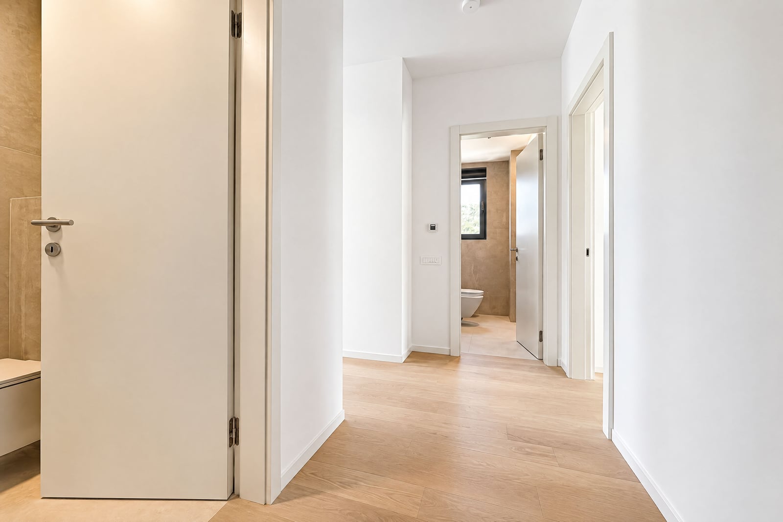 Bright hallway with light wood flooring and white walls, showing an open door to a bathroom with a toilet.