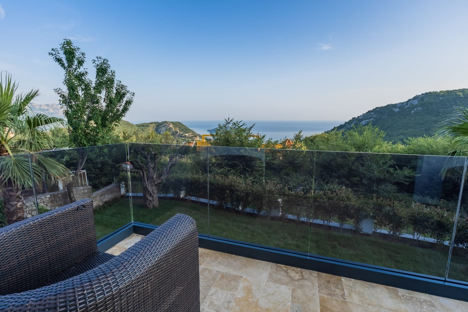 Villa terrace in Budva, Montenegro: wicker chair, glass railing, and panoramic view of the sea and mountains.