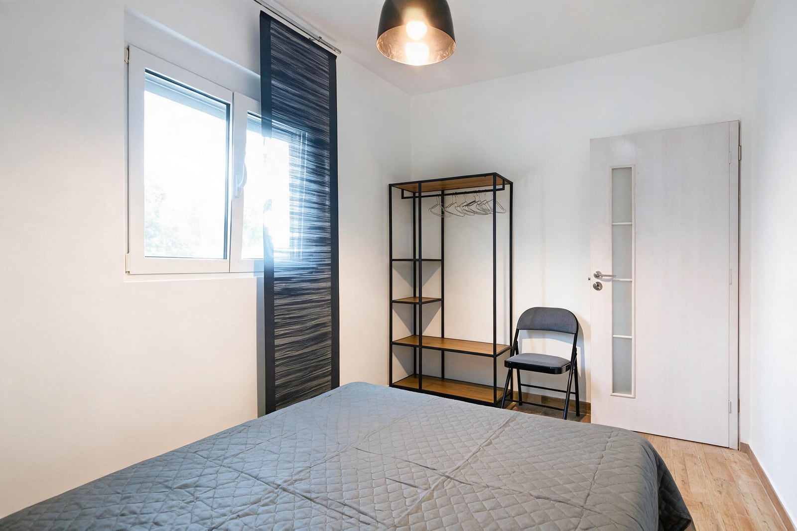 Bedroom featuring a bed with grey quilted cover, open metal wardrobe frame, folding chair, and window with dark curtain.