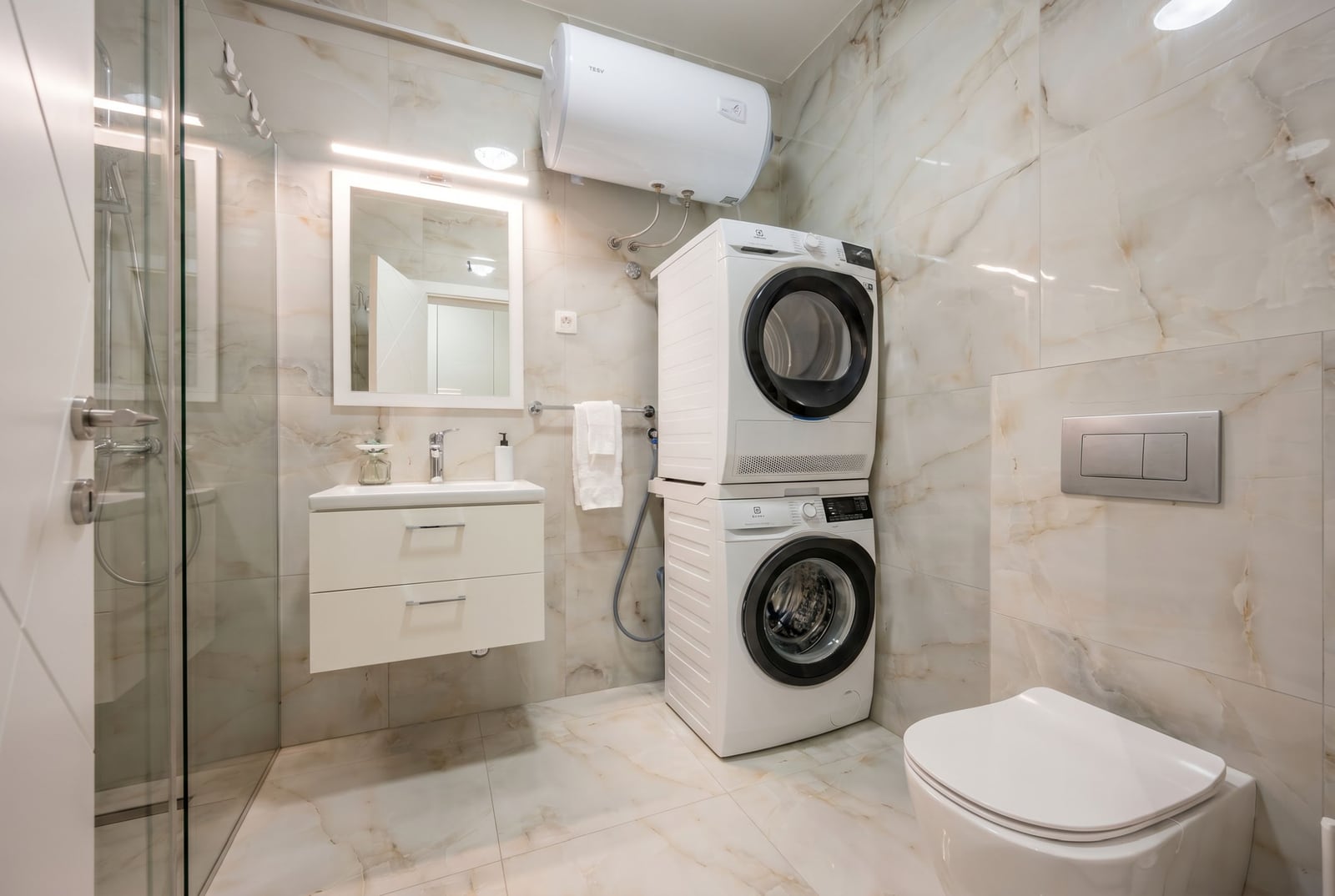 Modern bathroom in an apartment in Tivat, Montenegro: walk-in shower, washer and dryer, marble-effect tiles.