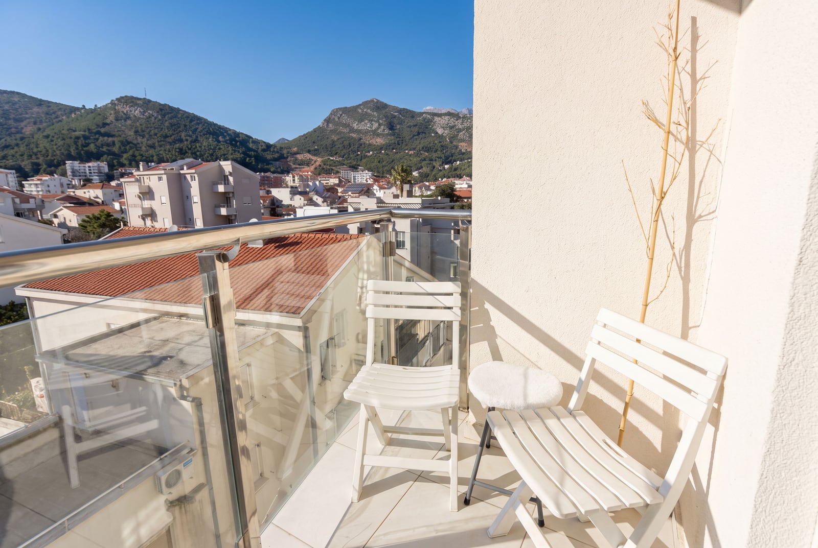 Balcony with white folding chairs and glass railing overlooking a town and mountains.