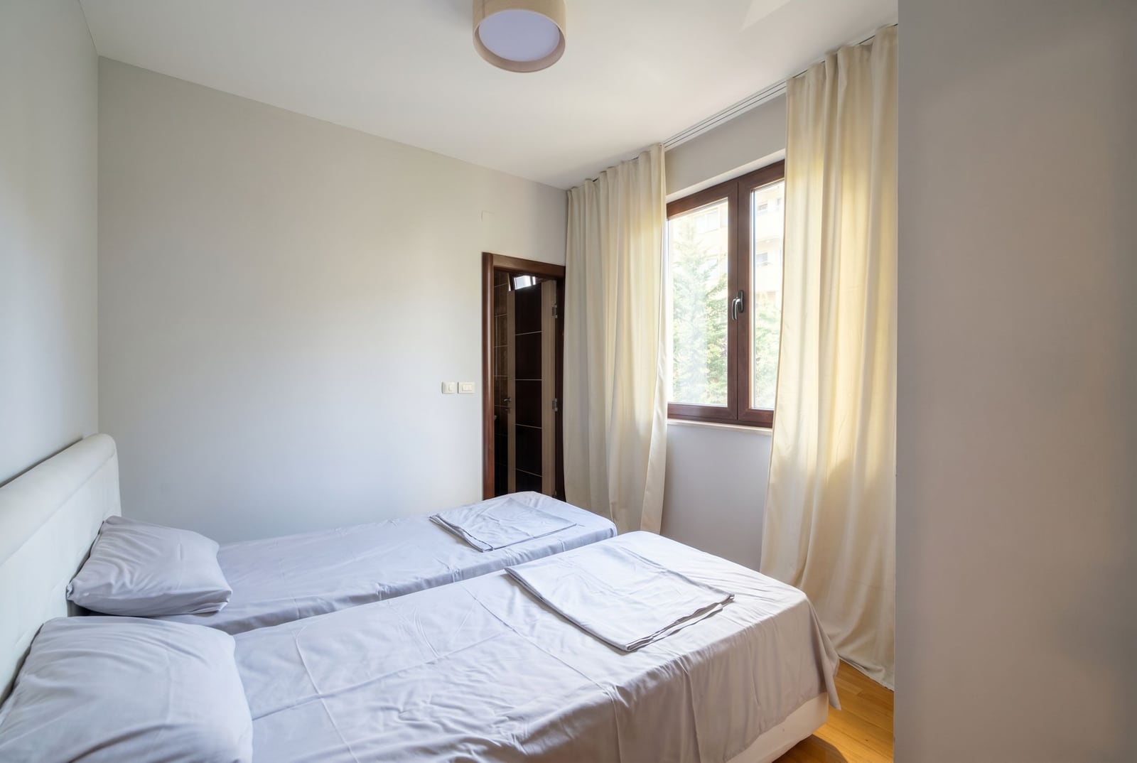 Bright bedroom with two beds in an apartment in Budva, Montenegro: modern interior, parquet flooring, and a view from the window.