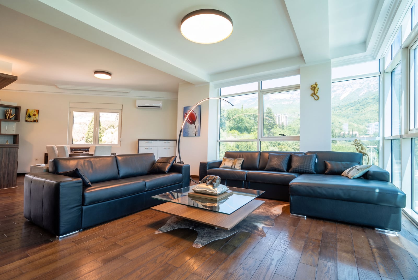 Bright living room with black leather sofas, a glass coffee table, and large panoramic windows.
