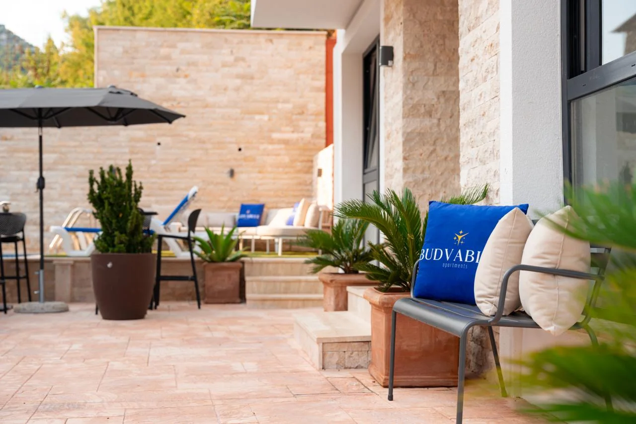 Open terrace with stone walls, outdoor furniture, umbrella, and potted plants next to a building entrance.