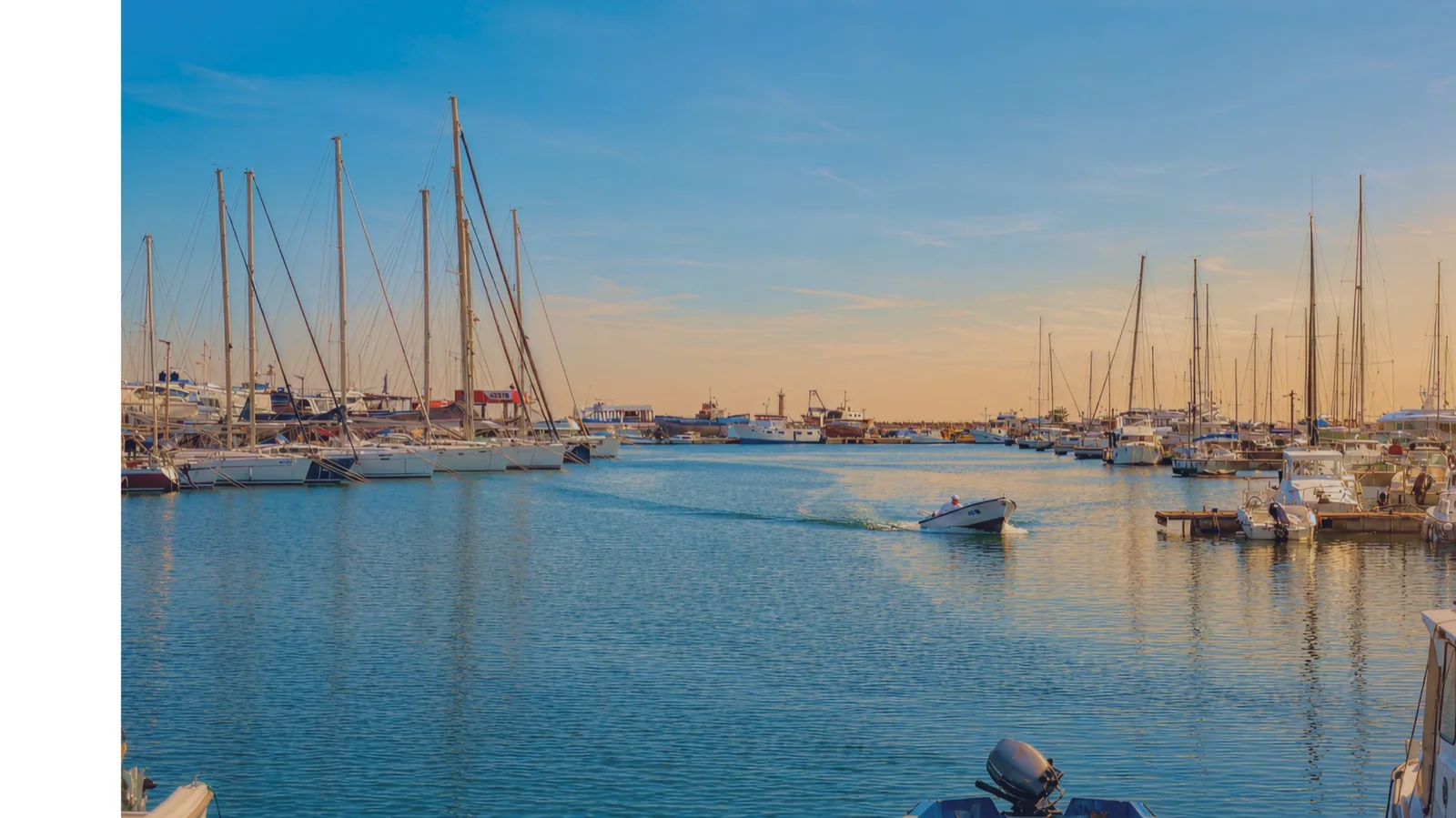 Bright photo of a marina filled with sailboats and motorboats on calm water under a clear blue sky during the day.