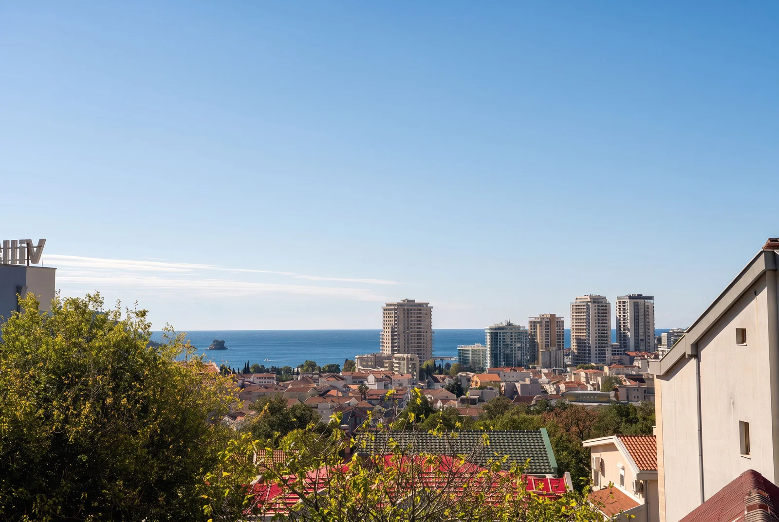Panoramic view of city rooftops, sea, and skyscrapers under a clear blue sky.