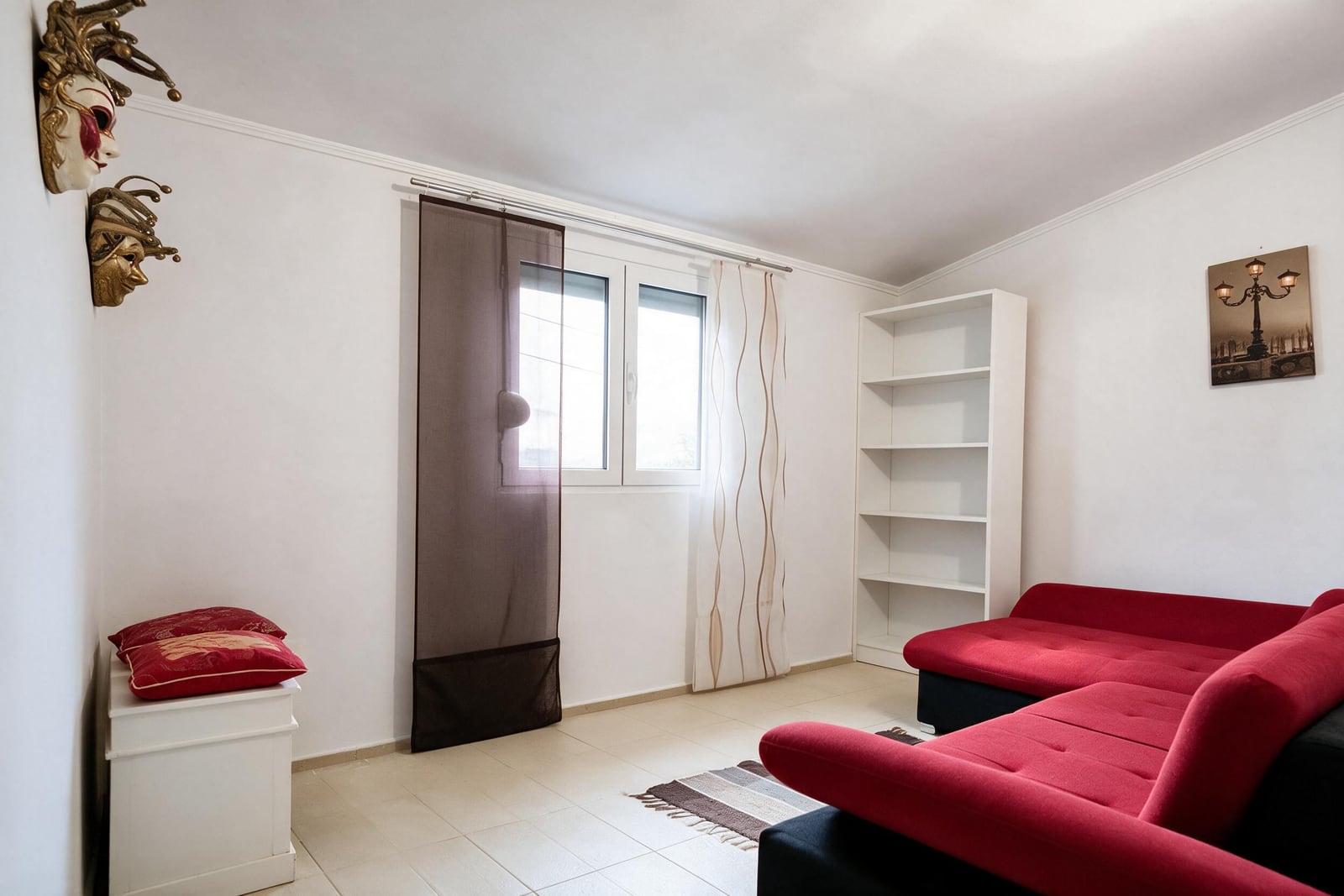 Living room featuring a red sofa, white bookshelf, decorative wall masks, window with curtains, and tiled floor.