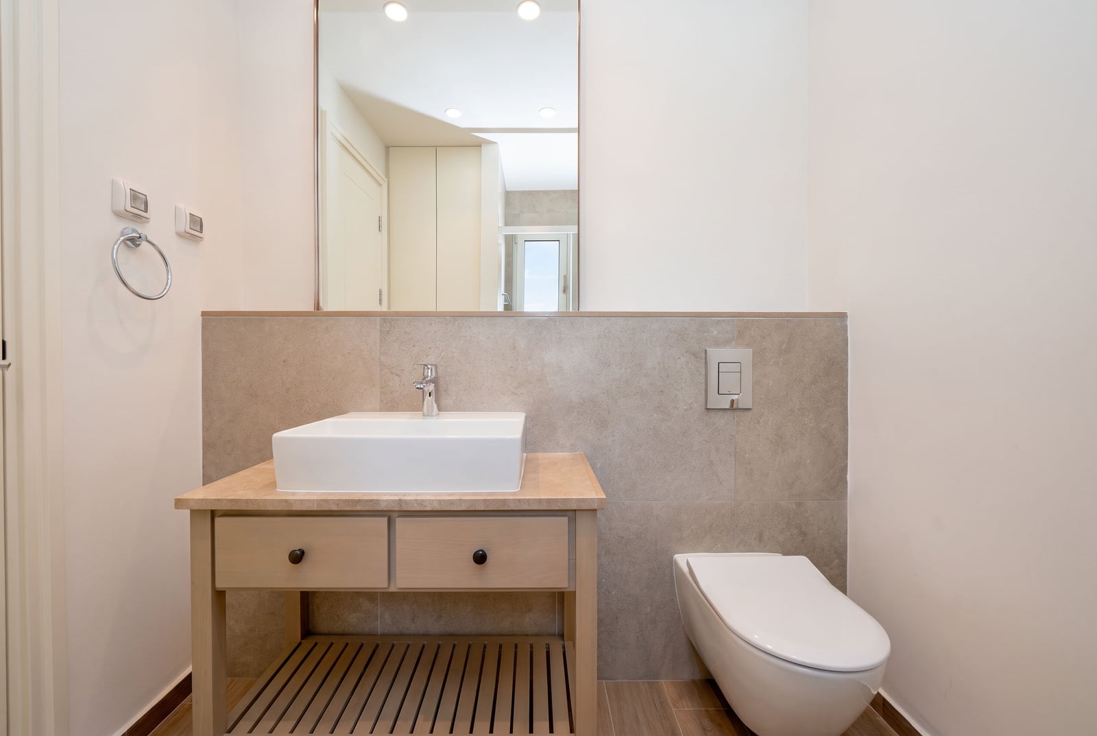 Modern bathroom featuring a wall-mounted toilet, wooden vanity with a rectangular vessel sink, and beige tiled backsplash.