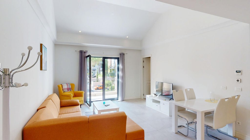 Bright living room with an orange sofa and dining area in an apartment in Luštica, Montenegro, with access to a balcony.