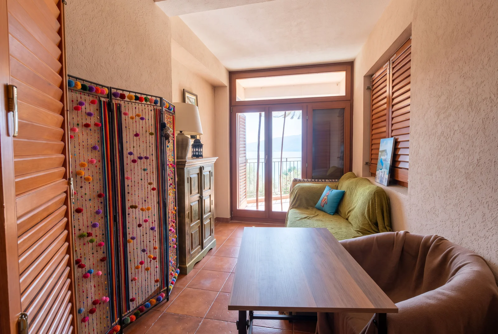 Living room with a wooden table, green sofa, decorative pom-pom screen, and glass doors leading to a sea-view balcony.