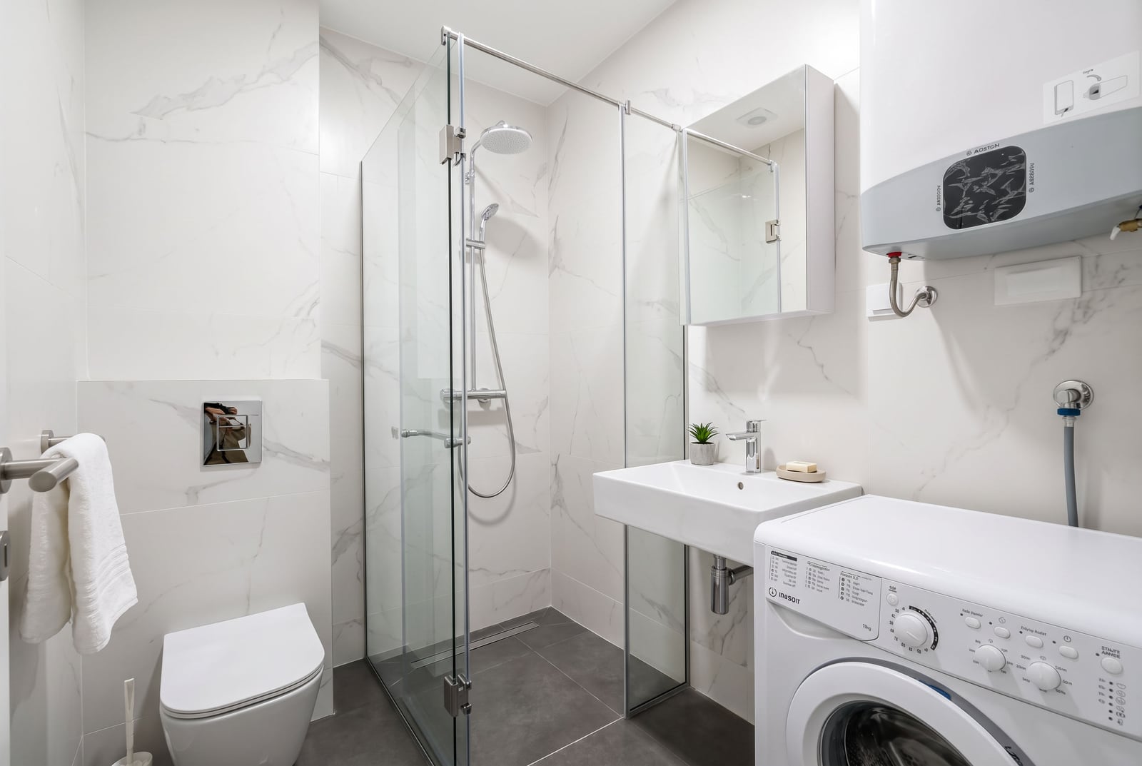 Modern bathroom featuring a glass shower enclosure, toilet, sink, washing machine, and wall-mounted water heater on marble tiles.