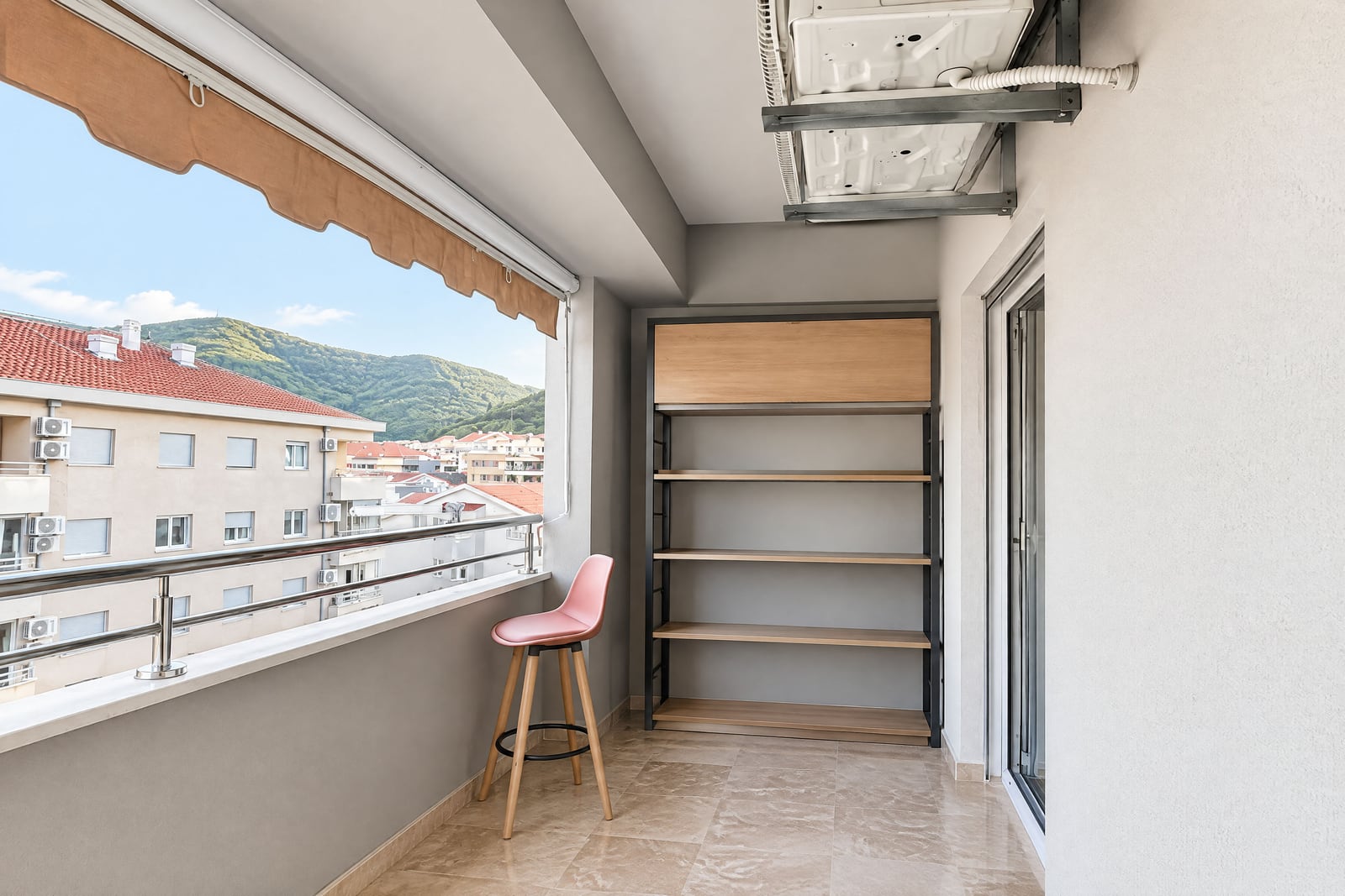 Balcony featuring a metal shelving unit, pink bar stool, retractable awning, and view of rooftops and hills.