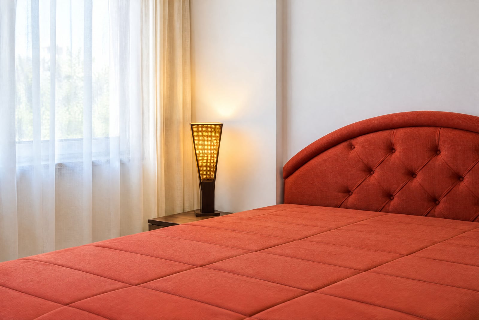Red bed with upholstered headboard and lamp on nightstand next to window with curtains.
