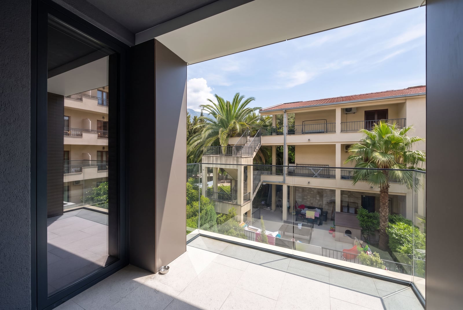 View from a spacious balcony with panoramic glazing overlooking the inner courtyard with palm trees and the residential complex in Budva, Montenegro.