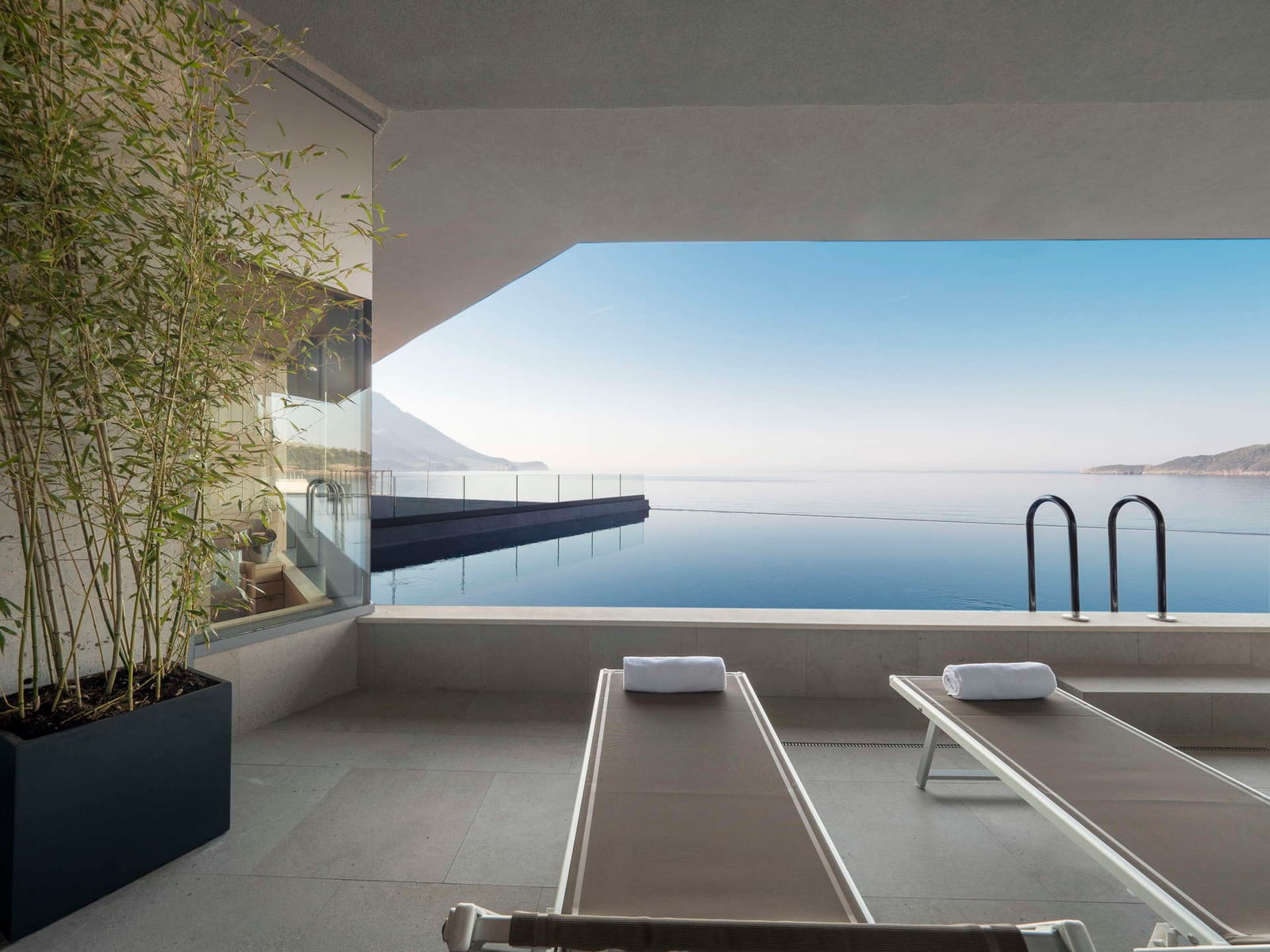 Infinity pool with sun loungers overlooking the sea, featuring potted bamboo and a glass barrier.