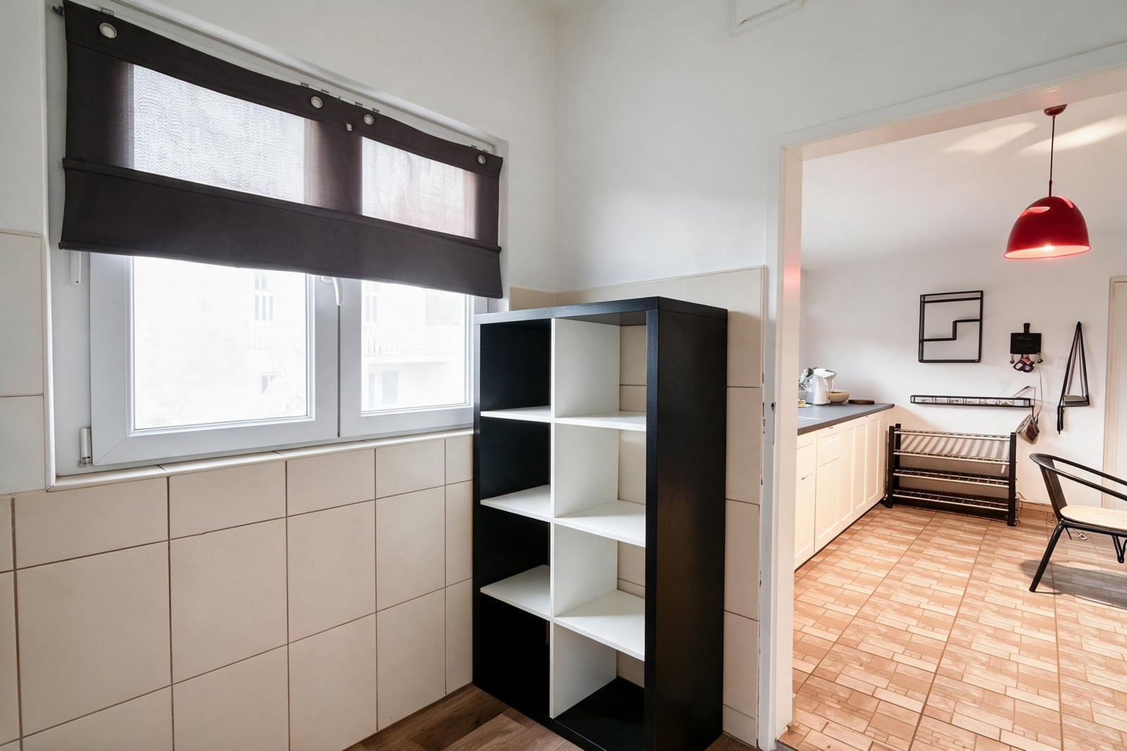 Dark shelving unit by a tiled window; adjacent room shows kitchen counter, red pendant light, and shoe rack.