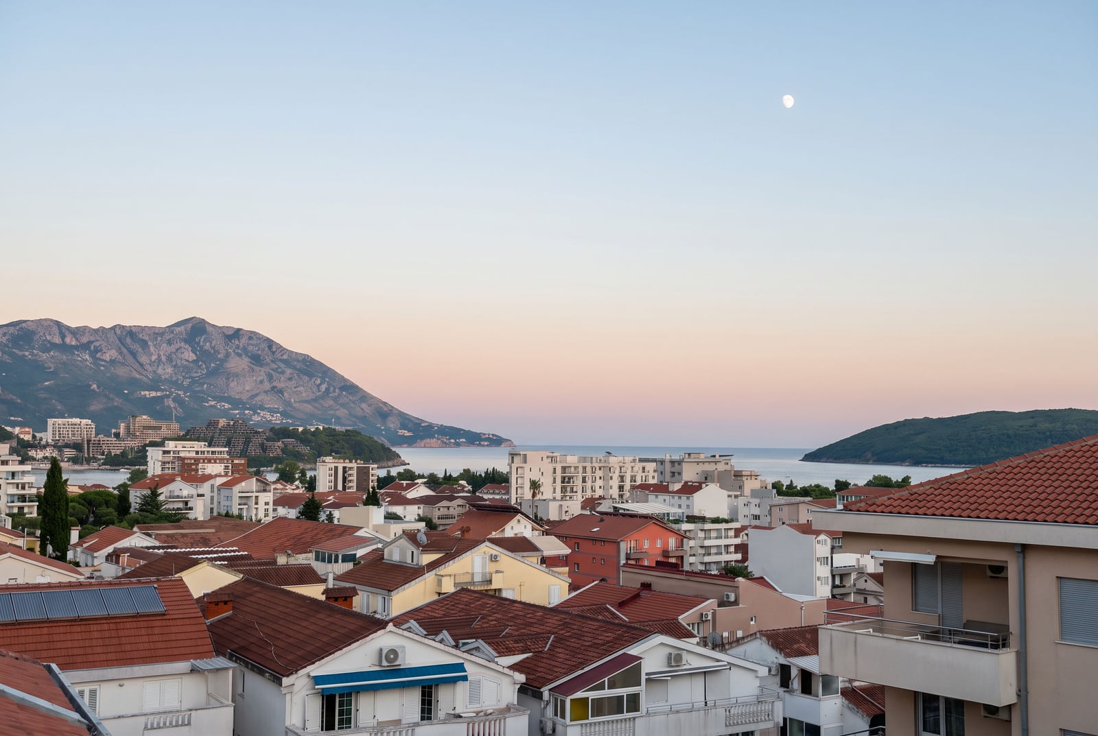Panoramic view of city rooftops, sea, and mountains against a sunset sky with the moon.