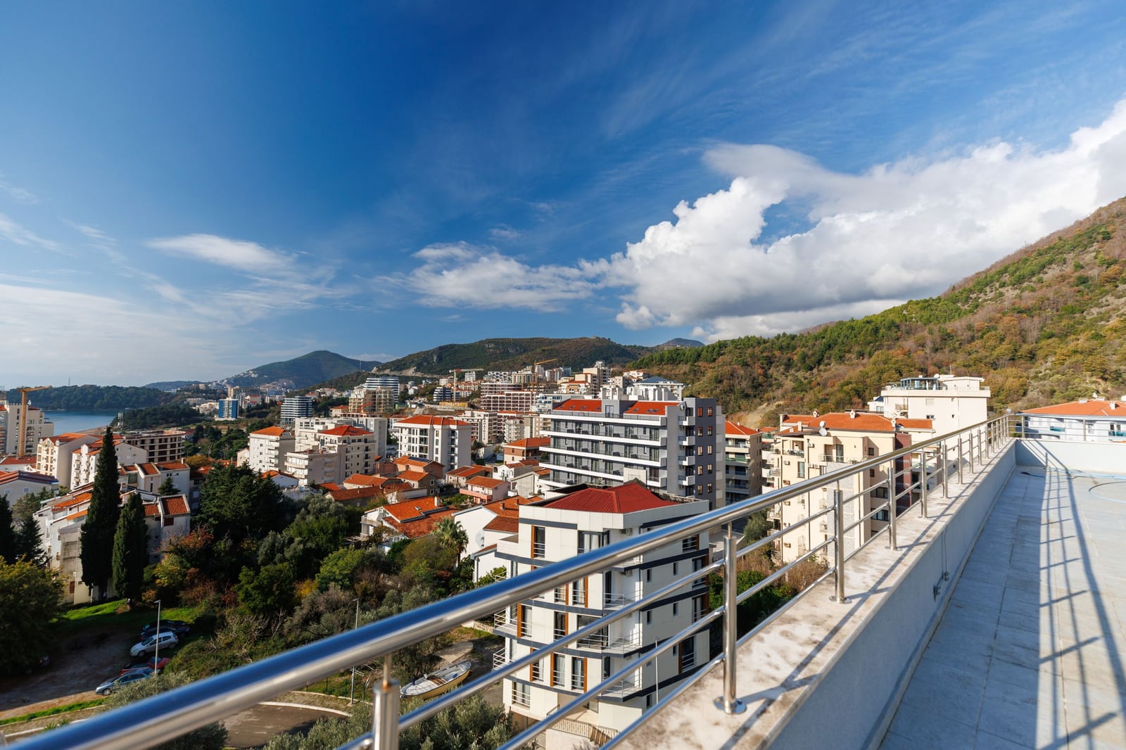 Panoramic view from the penthouse terrace in Bečići, Montenegro: city, mountains, and sea, with modern stainless steel railings.