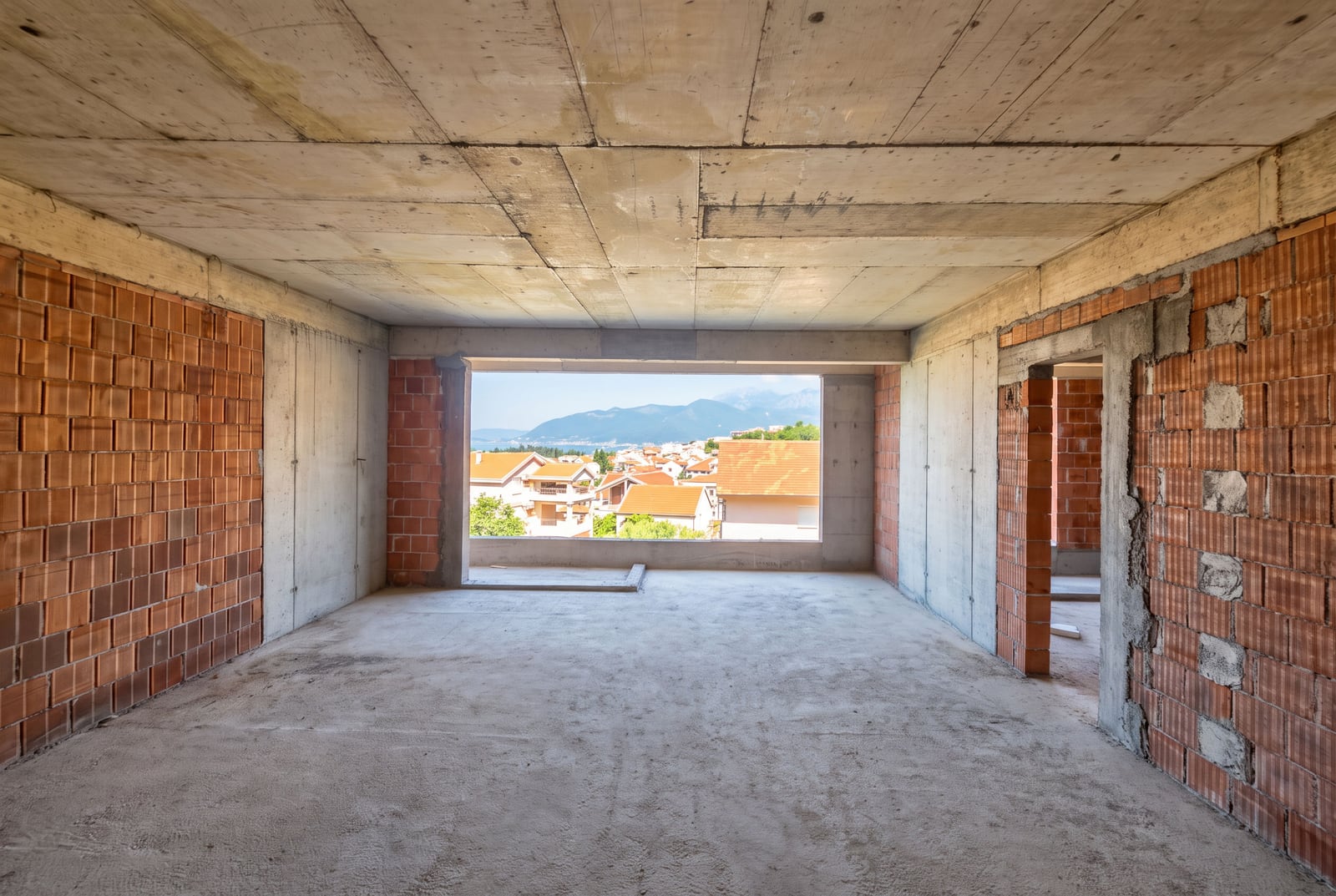 Unfinished room with concrete ceiling and brick walls, overlooking a town and mountains.
