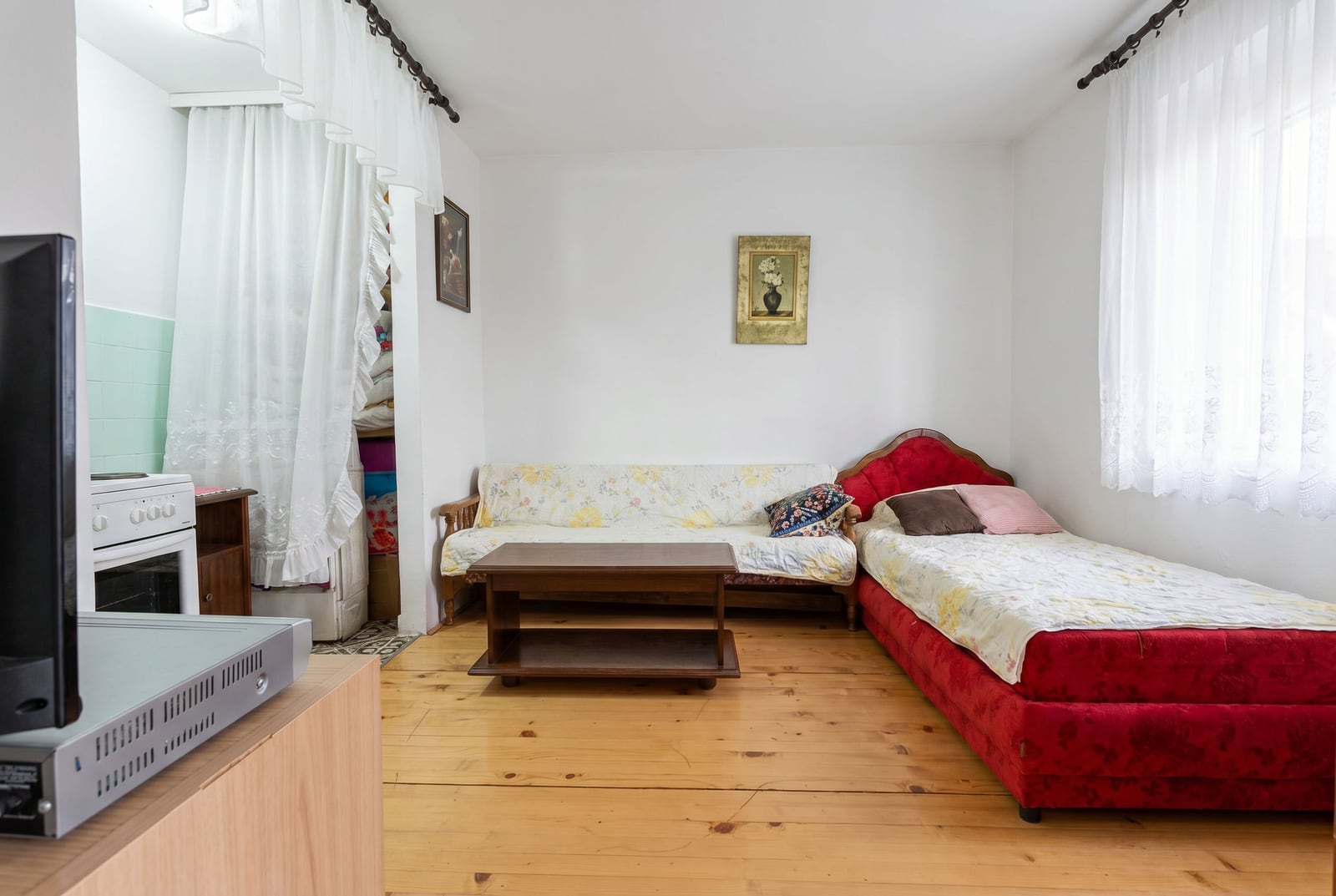 Living room in a house in Sutomore, Montenegro: wooden floor, red bed, sofa, kitchen area, and bright interior.