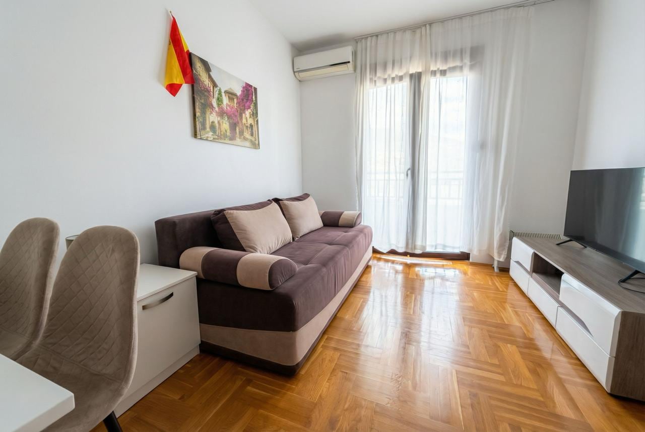 Living room in an apartment in Budva, Montenegro: brown sofa, parquet flooring, TV area, and access to a balcony with panoramic windows