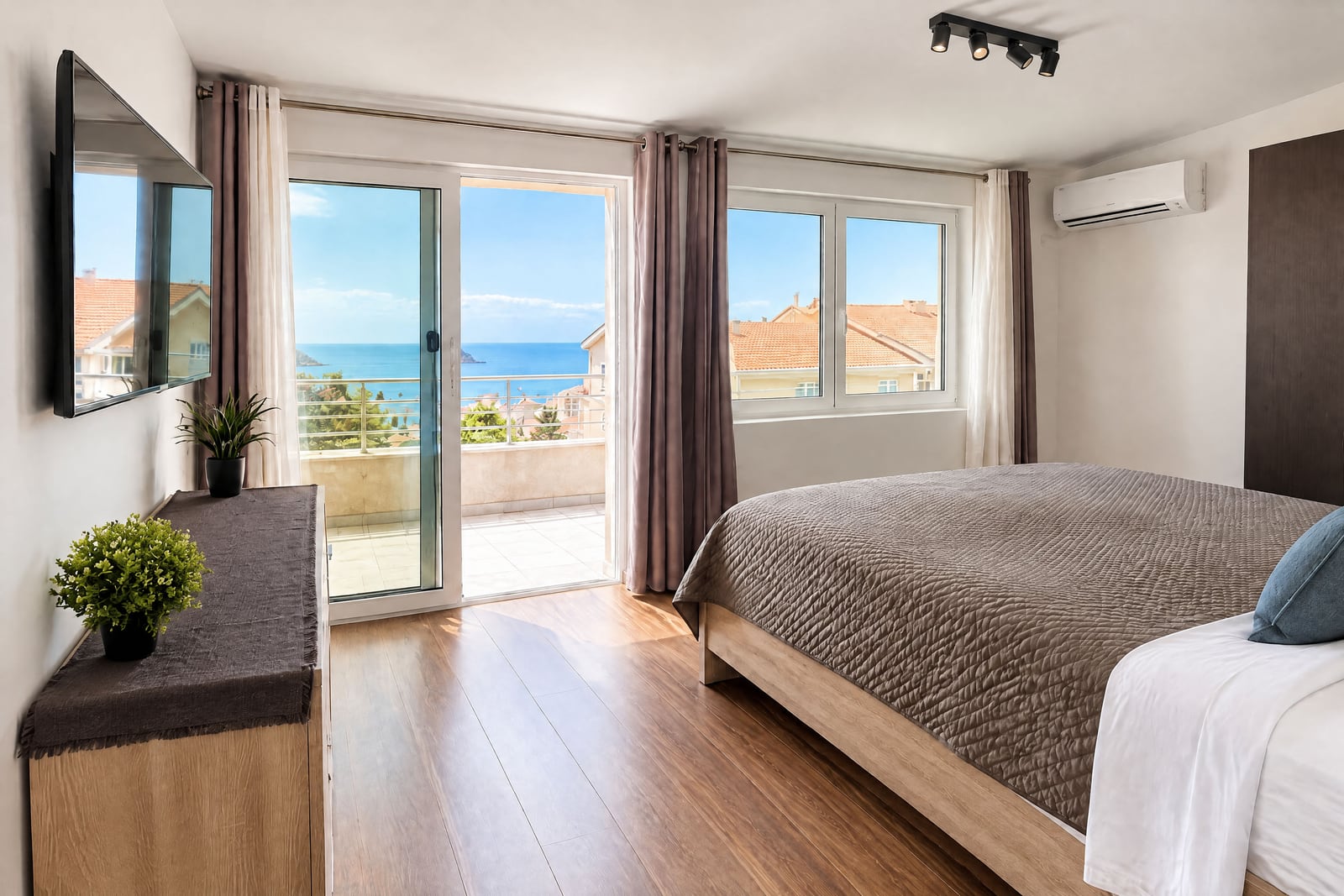 Modern bedroom with a double bed, wall-mounted TV, and sliding doors leading to a balcony overlooking the sea.