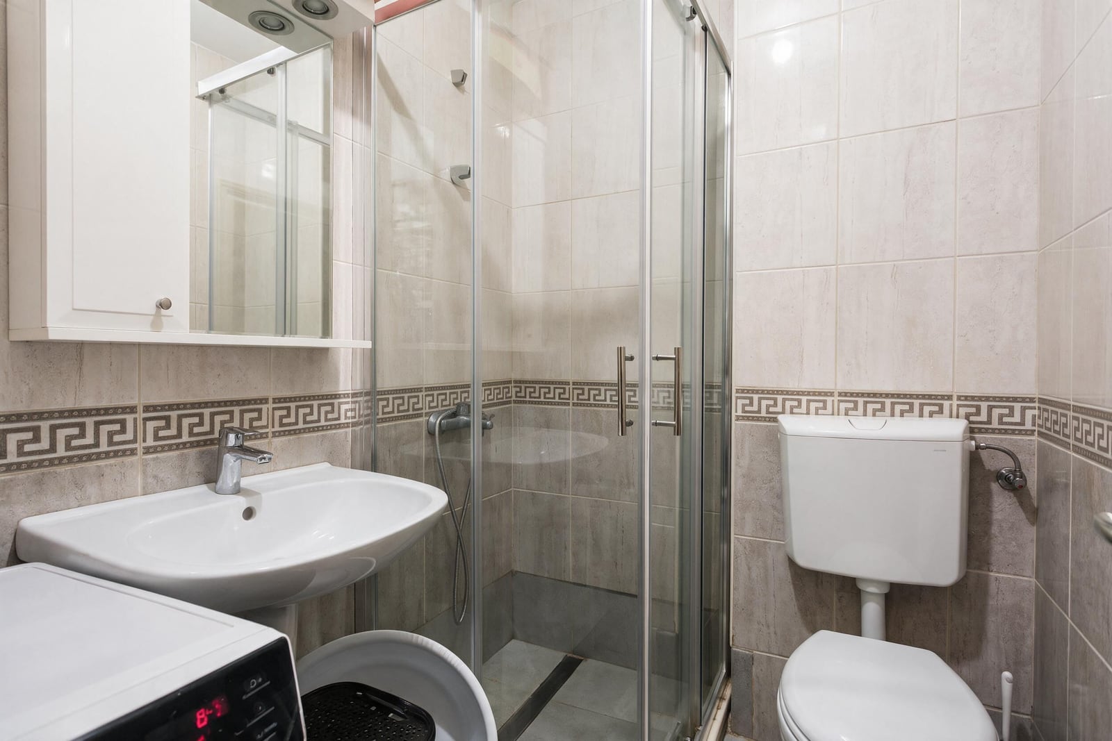Bathroom featuring a glass shower enclosure, white sink, toilet, and washing machine on tiled walls.