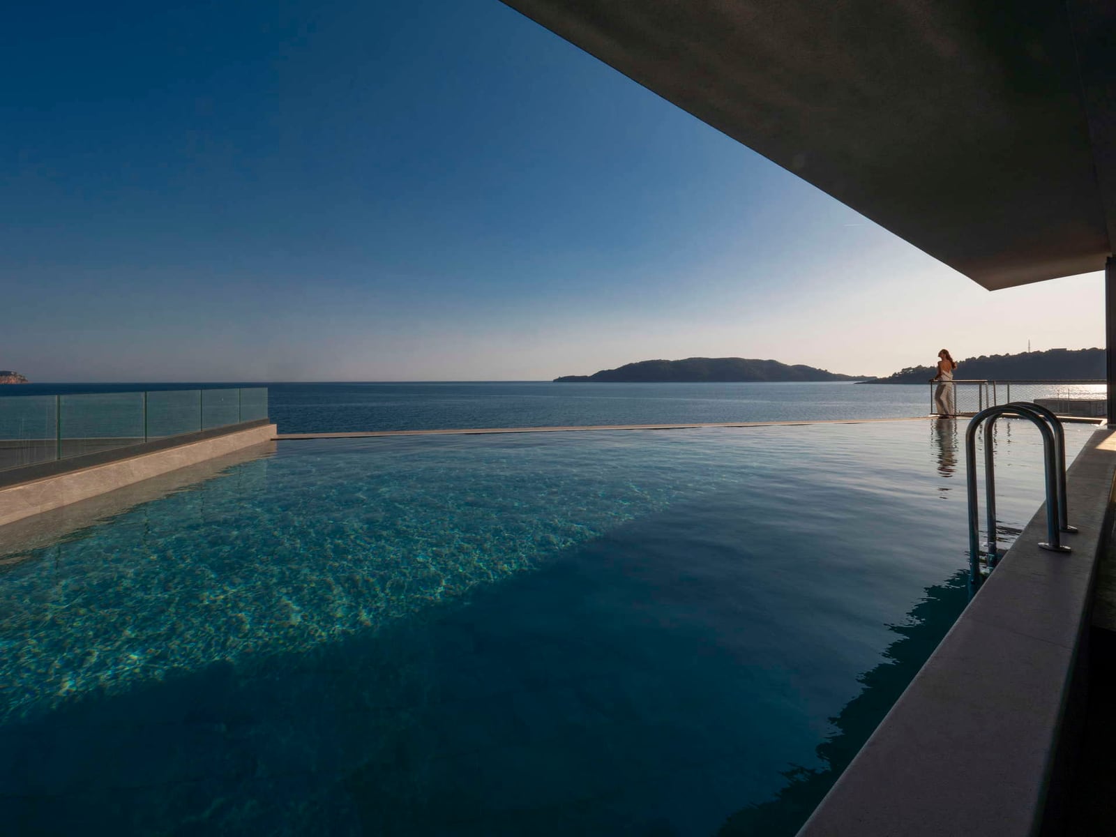Infinity pool overlooking the sea and an island. Metal ladder and glass railing are visible.