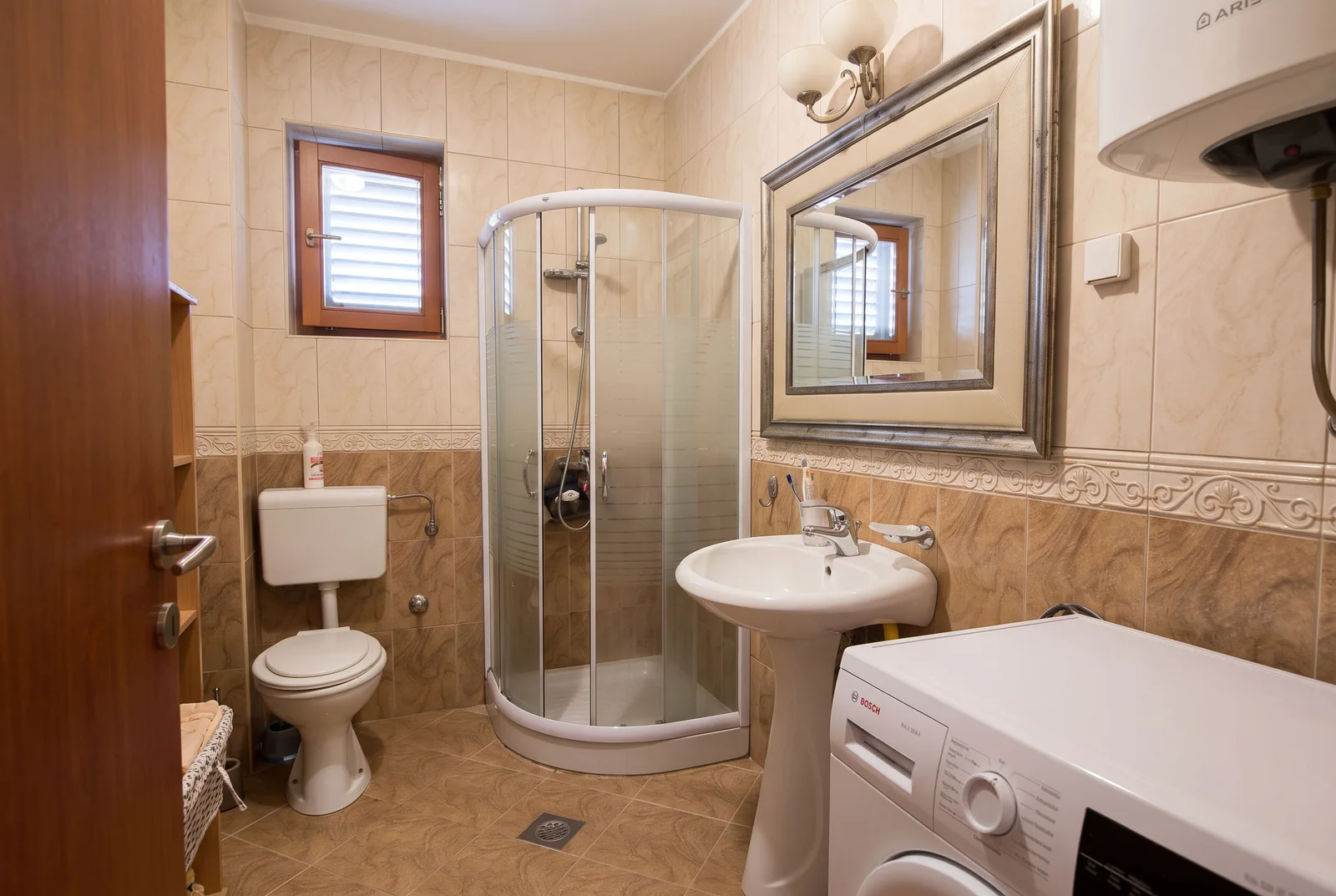 Bathroom featuring a shower stall, toilet, pedestal sink, and washing machine on beige tiles.