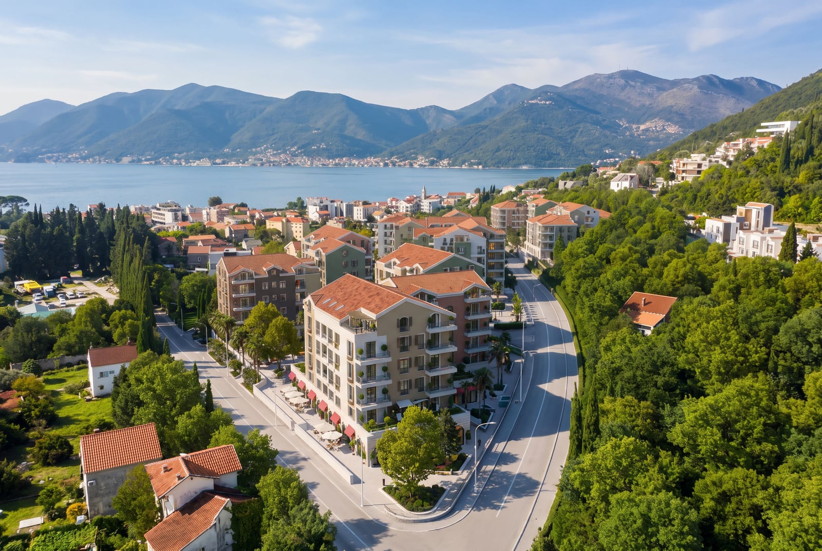 Aerial view of a coastal town featuring multi-story buildings, roads, lush greenery, sea, and mountains in the background.