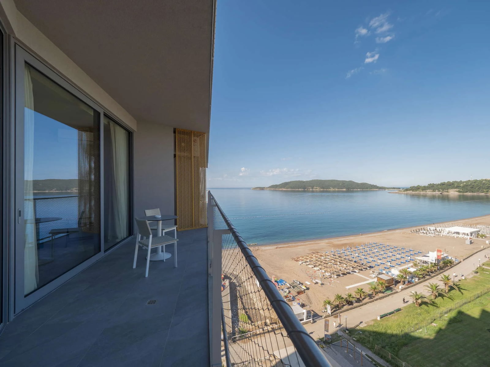 Balcony with white table and chairs overlooking a sandy beach and blue sea through a metal railing.