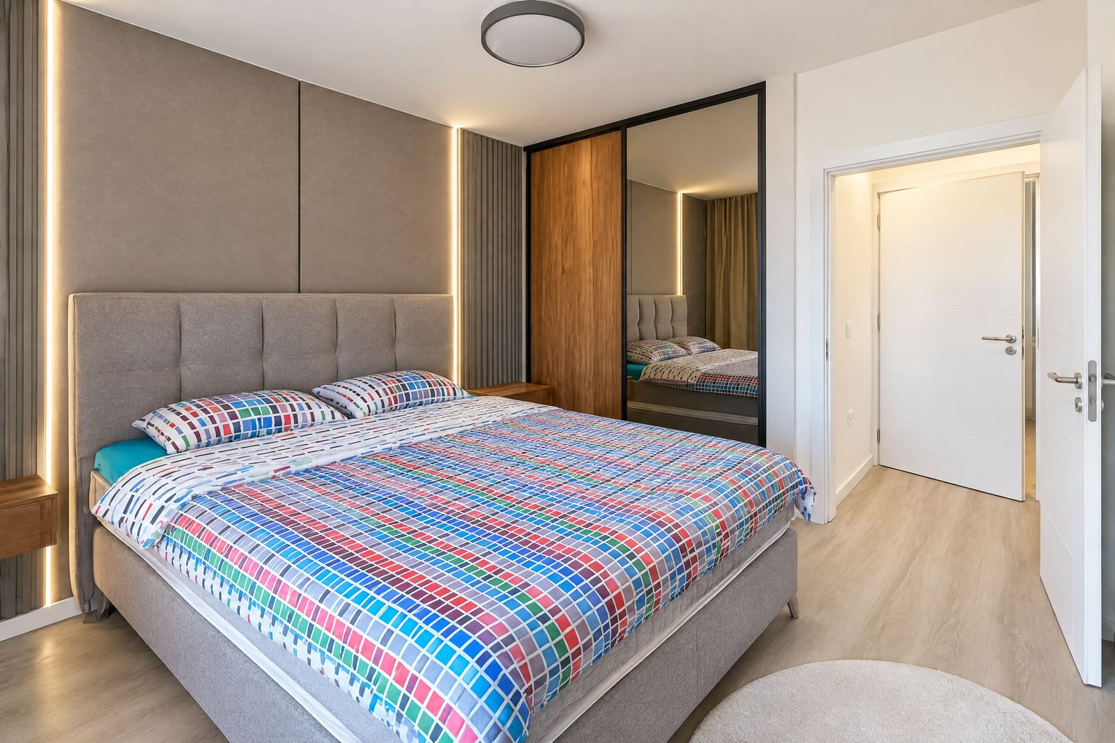 Modern bedroom featuring a double bed with colorful bedding, upholstered headboard, and mirrored wardrobe doors.