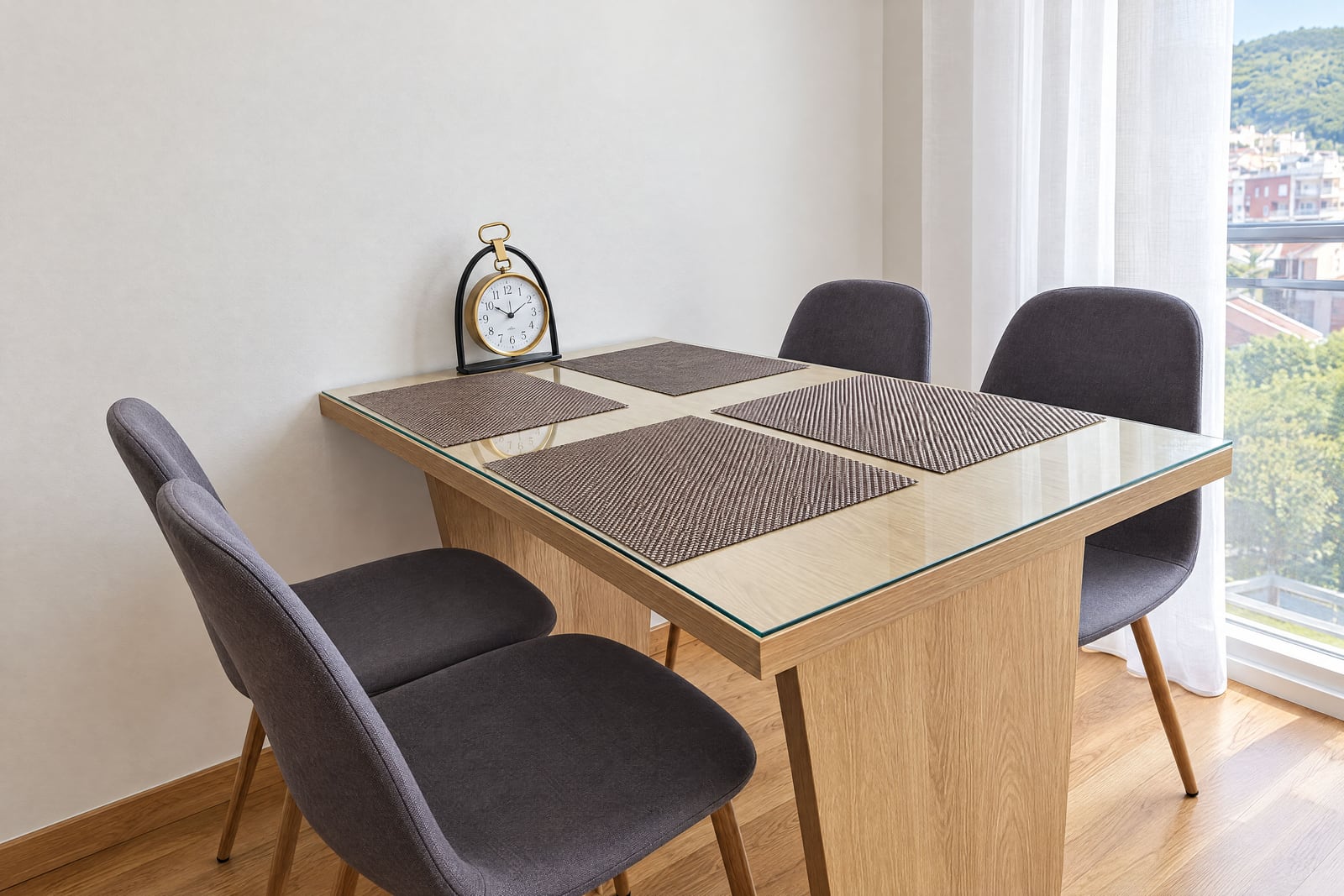 Bright dining area with a glass-top table, four grey chairs, and a desk clock near a window with sheer curtains.