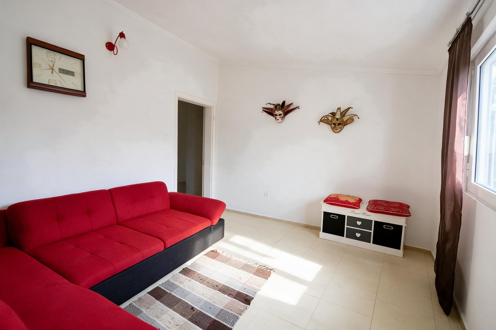 Red corner sofa, wall masks, clock and white storage cabinet in a room with light tiled flooring.