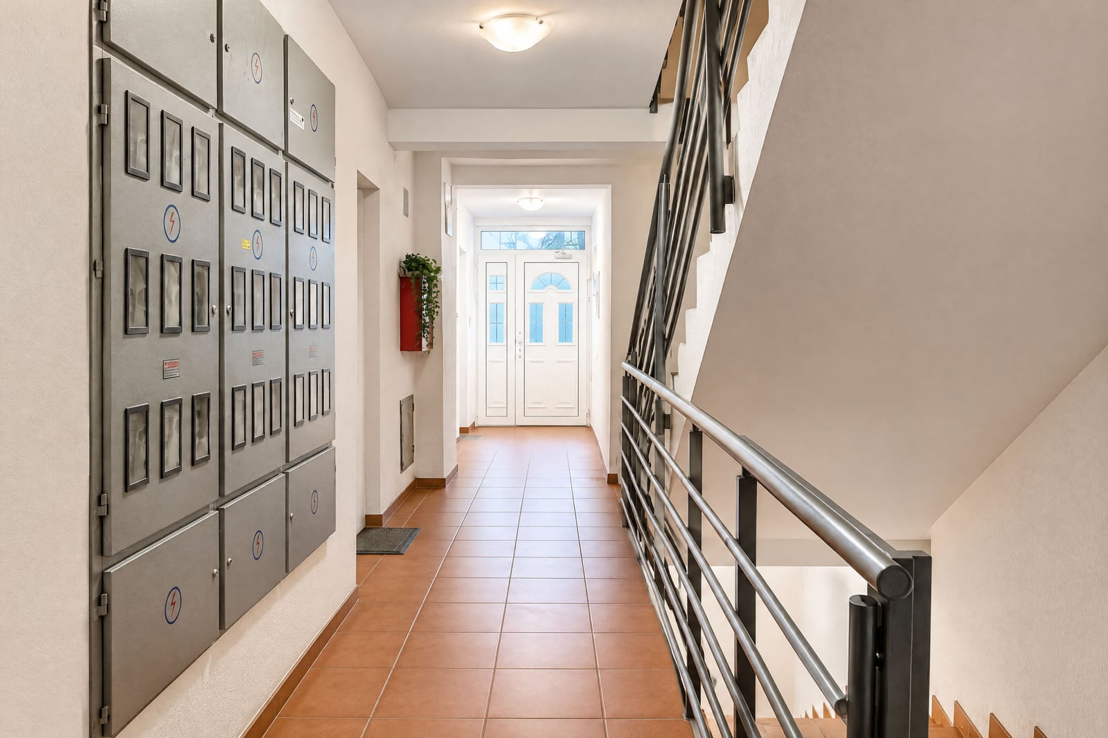 Hallway with electrical panels, staircase railing, and tiled floor leading to an entrance door.